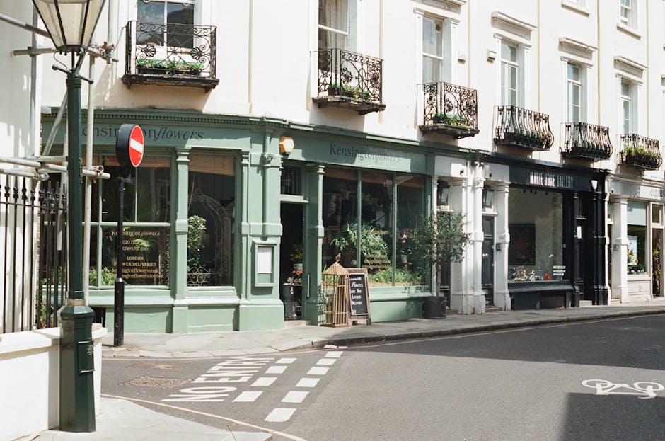 A storefront of a flower shop with large display windows filled with fresh floral arrangements, painted in a soft muted green color, situated on a street corner in Clerkenwell. The shop has an elegant