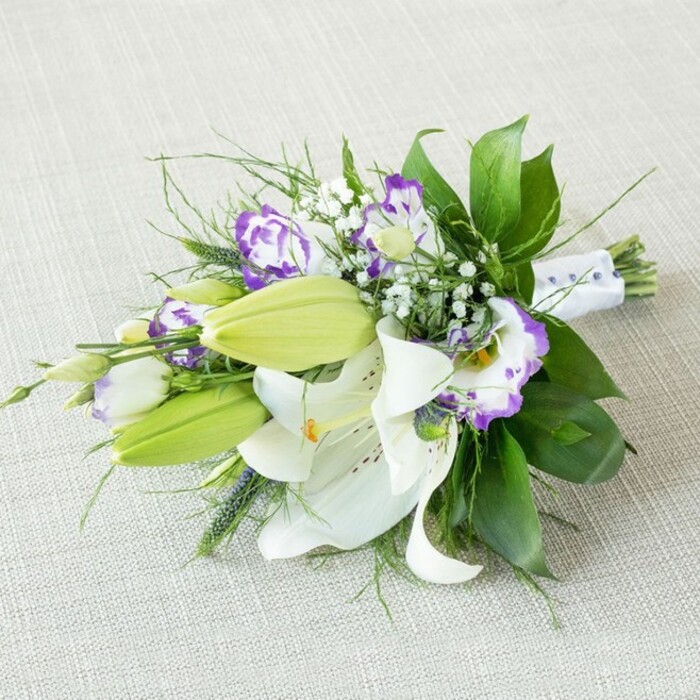 A refined, hand-tied bouquet stretches diagonally across a gently woven, light beige fabric, its elongated shape leading the eye from one end of the arrangement to the other, much like a floral ribbon laid out in a Clerkenwell studio. At the visual centre, several large Oriental lilies bloom in pristine white, their broad petals slightly recurved and satin-smooth, drawing attention to the prominent yellow-orange stamens and faint, rust-coloured speckles that cluster near each flower's throat. Around them, sleek, pale green buds stand upright, yet to unfurl, infusing the design with a sense of unfolding time and future celebrations. Interlaced among the lilies are numerous Lisianthus blossoms, their distinctive ruffled petals forming soft, cupped shapes. Each bloom transitions from creamy white bases to striking purple edges, some tinted with light lavender, others with deeper violet tones, creating a layered, almost watercolour effect. Sprays of delicate white Gypsophila are scattered throughout, like fine mist or a sprinkling of stars, filling gaps and adding gentle volume. The greenery is rich and varied: wide, glossy dark green leaves ground the composition, while finer, linear fronds and a few sculptural, spiky elements introduce texture and movement, evoking the mix of historic brick and contemporary design seen around Clerkenwell's workshops and galleries. At the base, the stems are neatly gathered and secured with a crisp white ribbon that wraps tightly, discreetly accented with small, pearl-like beads aligned along the narrower end, hinting at bridal craftsmanship from a local florist near Clerkenwell Road. Soft, diffused lighting glows across the bouquet, casting minimal shadows and allowing every petal, leaf vein, and bead to be clearly perceived. The image conveys freshness, sophistication, and a gentle romantic energy, fitting for wedding flowers, a special anniversary delivery, or an elegant centrepiece for a dinner just off Farringdon Lane.