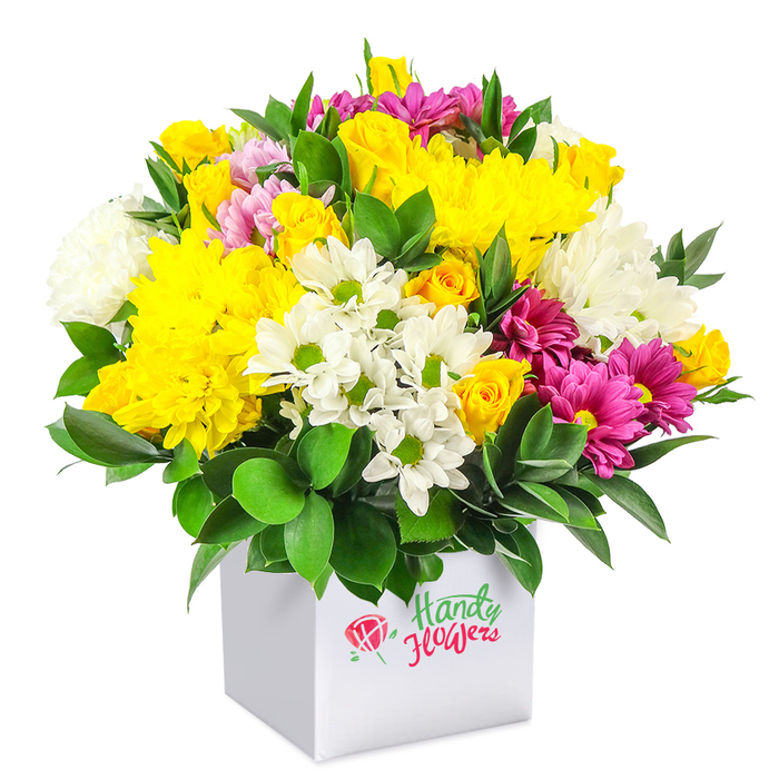 A lush and tightly composed bouquet is displayed in a modern white square box, set before a plain white backdrop that allows every bloom and leaf to stand out clearly. The arrangement forms a smooth, rounded dome, brimming with fresh flowers in sunny yellows, bright whites, and vibrant fuchsia tones, supported by a collar of deep green foliage. Dominating the design are large yellow chrysanthemums and clusters of yellow spray roses, some fully open and others still in bud, their velvety petals overlapping to create a soft, almost cloud-like texture. Interspersed throughout are white chrysanthemum sprays that resemble daisies, each with a vivid green centre, alongside fluffier white mums that introduce volume and a cooling brightness. Cutting through this gentle contrast, fuchsia and magenta chrysanthemums, some tinged with lighter pink, offer flashes of rich colour and give the bouquet a contemporary feel that would look at home in a Clerkenwell townhouse or office near Old Street. Around the edges and between the blossoms, glossy dark green leaves frame the composition, adding depth and a subtle shine that grounds the cheerful palette. The flowers sit snugly in a pristine cube gift box that doubles as a vase, its straight lines and clean surfaces enhancing the sculpted, dome-like profile of the blooms. This image conveys the care of an experienced local florist, the kind who understands how to arrange something joyful yet refined for everyday celebrations across Clerkenwell - from birthday breakfasts in converted warehouse apartments to thank-you gestures delivered to creative studios and co-working spaces nearby.