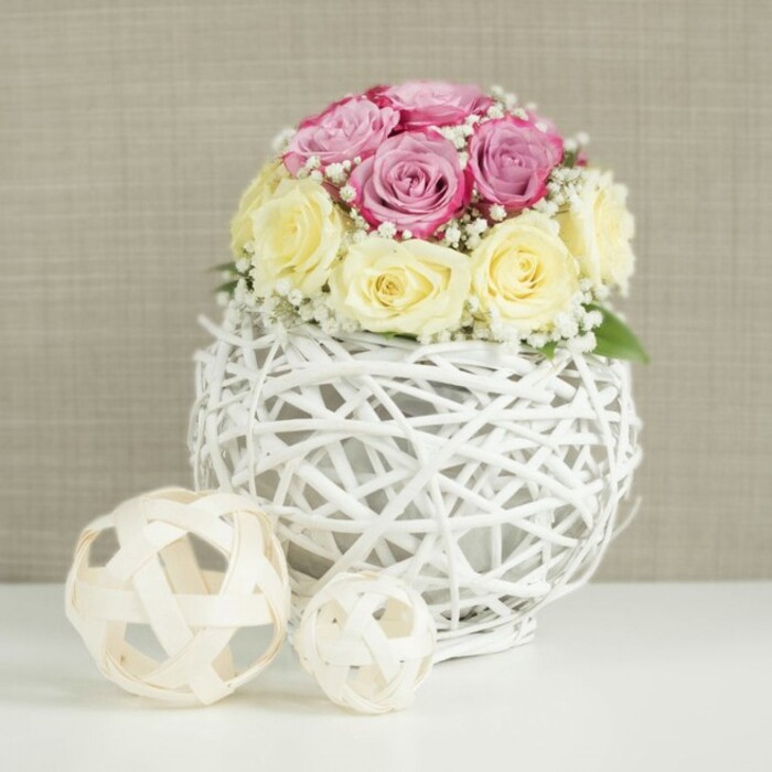 A delicately composed floral centerpiece is shown in close view, perfect for a wedding or intimate dinner party in Clerkenwell's converted warehouses and townhouses. At the heart of the scene, a compact, rounded bouquet of roses sits in a spherical white wicker vase, its woven strands forming an intricate openwork cage. The roses number around a dozen and are balanced between two soft, romantic tones: half in a gentle, powdery pink with a touch of fuchsia warmth, and half in a creamy yellow that lightens to ivory at the petal edges. Some blooms are just beginning to open, their petals cupped and tight, while others spiral outward in lush, velvety layers, giving the dome a pleasing mix of structure and softness. Interlaced throughout are fine stems of white baby's breath, their tiny, cloud-like blossoms filling the spaces between the roses, creating a light, airy veil that softens the overall look the way early evening light softens the streets around St John's Square. The bouquet is cradled in a distinctive white-painted wicker globe, its thin, criss-crossed branches creating an airy lattice that reveals hints of the inner container and casts gentle shadows onto the white surface below. To the left, two decorative spheres crafted from pale natural wood strips repeat the woven motif, one larger and one smaller, resting casually on the same clean, white tabletop. Behind the arrangement, a neutral beige, linen-like wall provides a warm, understated backdrop that highlights the pastel tones without distraction. The mood is calm, elegant, and welcoming, well-suited to a Clerkenwell Flowers table design for a small wedding reception, engagement celebration, or stylish home gathering near Farringdon or Hatton Garden.