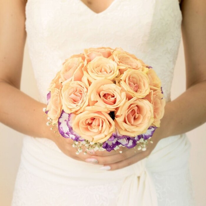 The photograph focuses on a serene bridal moment, framed from the chest down so that the viewer's attention naturally rests on the bouquet and the refined details of the gown. The bride stands against a softly blurred, light-toned background, reminiscent of a bright studio space or a sunlit room off Farringdon Road. Her dress is a traditional white or ivory, with a delicately structured lace bodice that forms a gentle V-neckline. The lace is fine and romantic, suggesting tiny blossoms and leaves, while the skirt below falls in smooth, slightly folded fabric that hints at movement and grace without cluttering the scene. Held carefully in both hands is a rounded bridal bouquet, clearly the work of a skilled florist used to styling weddings around Clerkenwell's intimate venues and historic churches. The upper section of the bouquet is formed from an abundance of roses in soft, glowing tones of peach, apricot, light orange, and blush pink. Their petals are tightly layered and slightly cupped, each bloom pressed close to its neighbours to create a neat, domed silhouette that feels both lush and controlled. Beneath these roses, a rim of ruffled flowers in rich purple with creamy white edges-likely lisianthus-creates a striking collar, giving the bouquet depth and visual interest. White baby's breath is interwoven around the base, adding a fine, cloud-like texture that softens the transition between colours. The stems are gathered compactly, disappearing behind the bride's lightly tanned hands. Her nails are trimmed and polished in a soft nude shade, with one thumb catching the light in a subtle ombré or glitter effect that echoes the bouquet's gentle sparkle. The overall impression is of romantic sophistication and thoughtful design, perfectly suited to a modern London wedding that values both tradition and individuality.