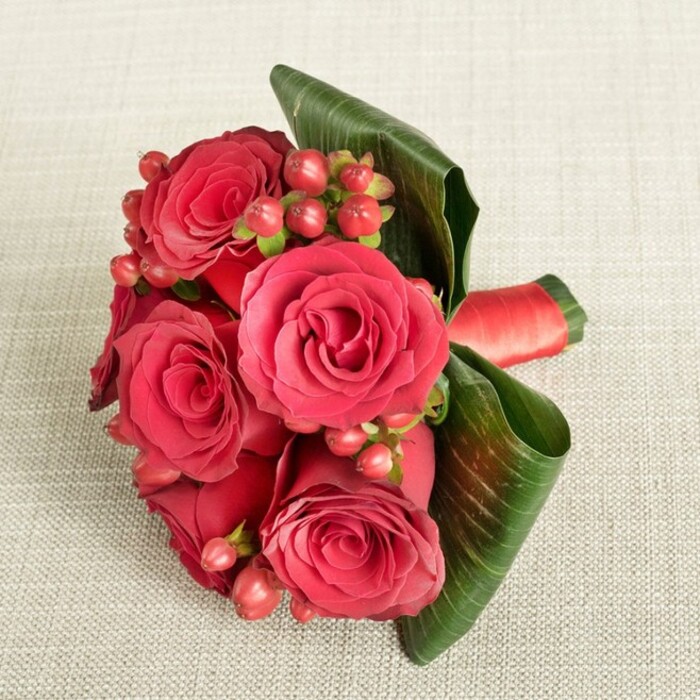 A compact, luxurious bouquet dominated by deep red roses rests on a pale beige, finely textured fabric, the neutral background softly diffusing the light and allowing every petal and berry to stand out with clarity. The roses are at their peak, petals layered densely from tight, shadowed centres to outer whorls that curl back with a velvety softness, moving from almost burgundy depths to brighter crimson edges. Between these plush blooms, clusters of round Hypericum berries in deep red and flushed pink nestle in, their smooth, bead-like forms providing a playful yet refined textural contrast. Slender green stems thread discreetly through the composition, barely visible but adding a sense of natural growth. Encircling the base, broad, dark green leaves are deliberately shaped and curled, forming a firm, glossy collar that cradles the roses and berries like a frame around a painting. The veins of the leaves catch the light subtly, suggesting resilience and freshness, as if just prepared in a local studio off Clerkenwell Road. A rich red satin ribbon binds the stems, hidden beneath but peeping out at one side with a gentle sheen, echoing the colour of the flowers and hinting at the florist's meticulous hand. Gentle, even lighting casts only the softest of shadows, enhancing the sense of depth without overwhelming the forms, and creating a mood of quiet sophistication. This bouquet feels perfect for an anniversary dinner near Smithfield Market, a Valentine's gesture sent across EC1, or an intimate wedding detail, its classic colour palette and careful craftsmanship conveying enduring love and elegant London style.