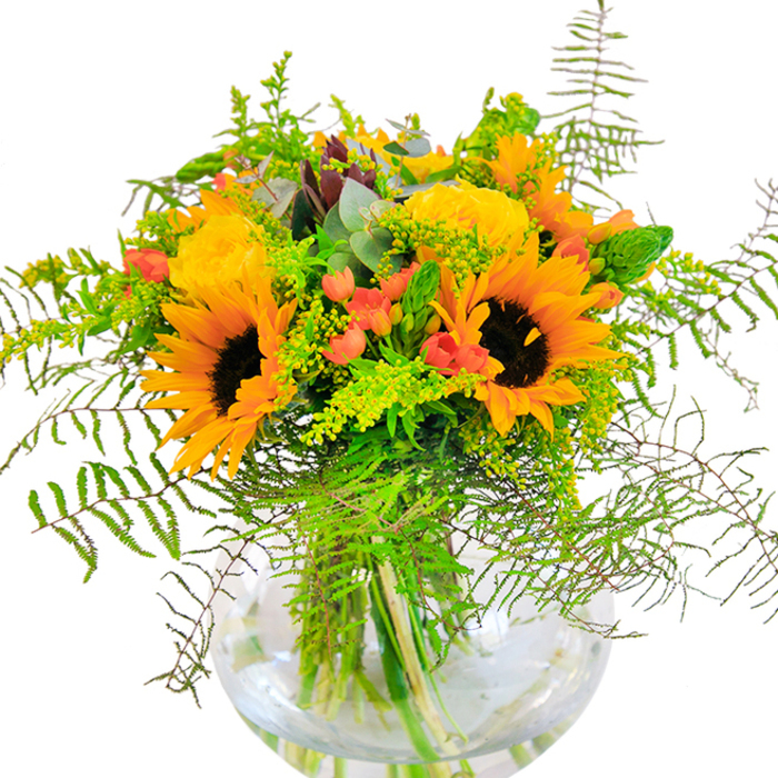 With the vibrant charm of Clerkenwell's flower stalls, this sun-inspired arrangement offers a vivid display of yellow sunflowers and roses, their golden hues radiating warmth and optimism. The bouquet's composition feels lush yet artfully balanced, anchored by large sunflowers whose sunset petals and inky brown eyes command attention. Between these, layers of yellow garden roses unfurl in spirals of plush texture, reminiscent of blooms freshly gathered from Leather Lane Market. Among the sunny tones, deep green leaves, airy vines, and slender spikes give the arrangement a gently cascading shape, softened by the feathery presence of fern fronds. Scarlet-red hypericum berries shine out, their glossy skins shimmering against the velvety petals and matte greenery. Wisps of eucalyptus leaves-tinged with subtle blue-green-peek between stems, providing aromatic freshness. Housed in a round, clear glass vase filled with glistening water, each stem is showcased in tidy elegance. The bouquet seems to catch natural morning light streaming into a Clerkenwell townhouse, creating luminous contrasts and soft shadows across the table. There's a delicate interplay of textures: sunflowers' suede, roses' silken petals, crisp foliage. With its gentle fragrance and exuberant visual energy, this arrangement is a perfect gift for moments of joy-a promotion, a birthday, or simply to bring a burst of local colour to a friend's day in EC1.