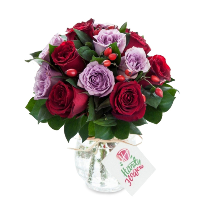 A richly coloured, skillfully composed bouquet centered on an even mix of deep red and soft lavender roses, arranged into a compact, rounded silhouette that reads as both luxurious and approachable. The reds vary from crimson to velvety burgundy; the purples move through lilac and dusty mauve, each bloom fully opened with visible unfurling petals that display their texture and depth. Scattered through the composition are small, bright red-orange rose hips that punctuate the arrangement with sparkles of autumnal colour and a rustic, tactile contrast to the smooth rose petals. Dark green foliage - likely Ruscus or Salal - is woven throughout, its elongated leaves creating pockets of shadow and visual breathing room between the flowers.

The flowers are presented in a bulbous, slightly tapered glass vase with a distinctive mercury or crackled silver finish; its reflective surface softens the light and lends a timeless, antique charm. A simple piece of natural twine is tied around the vase's neck, from which hangs a rectangular white gift tag illustrated with a stylised rose and neat green script, offering a handcrafted, personal touch. Photographed against pristine white, the bouquet's rich tones and the vase's muted gleam are emphasised, suggesting freshness and professional craftsmanship.

Prepared by Clerkenwell Flowers with local sensibility, this arrangement suits romantic gestures, anniversary surprises, or elegant home styling - the kind of bouquet you might pick up after a stroll past Exmouth Market or order for same-day delivery around Clerkenwell and Farringdon when you want something that feels both classic and quietly modern.