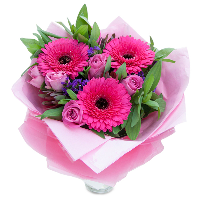 I like to think of this arrangement as the playful, graceful side of pink: three exuberant Gerbera daisies in a saturated fuchsia open wide, their layered petals fanning around a dark, defining centre that gives them striking presence. Surrounding these focal blooms, roses in a muted lavender-pink or dusty-rose unfurl with soft, rounded curves, each petal catching light differently and creating a sense of movement. Tiny clusters of purple-blue statice are tucked in like little surprises, their papery texture and cool hue adding depth and a meadow-like charm. The foliage - broad, textured leaves suggestive of salal and sprigs of eucalyptus - provides a fresh green frame, its glossy and matte surfaces forming shadow and relief. The bouquet is thoughtfully wrapped: an inner sheet of matte pale-pink tissue provides a gentle base while a layer of slightly crinkled, translucent fuchsia-pink cellophane overlays the flowers, giving a subtle sheen and reinforcing the colour theme. The top rim of a clear glass vase is visible below the wrap, hinting that the arrangement has been made to sit on a mantelpiece or bedside table at home. In cool morning light you might notice the roses' faint perfume and the gerberas' green-tinged sweetness; the textures invite a hand to linger. This composition is ideal for occasions that call for warmth without overstating it - birthdays, anniversaries, or a thoughtful surprise for someone returning to a flat near Spa Fields or shopping along Leather Lane - a little packet of care and cheer from a local florist's touch.