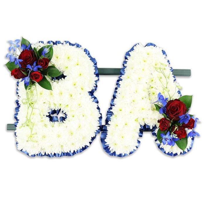 A carefully composed, symbolic floral tribute forming the uppercase initials BA, designed to read clearly from a distance yet reward close inspection with delicate detail. Each letter is completely covered in a dense field of pristine white chrysanthemum florets, their tiny petals overlapping to create a cushiony, matte surface that diffuses light and suggests peace. The perimeter of both letters is finished with a rich royal blue satin ribbon, ruffled and hand-placed to create a strong, elegant outline against the white. The B carries an accent cluster on its upper left curve of rich, velvety deep red roses, presented in varied stages of bloom so the eye moves from tight bud to open petal; these are combined with upright, electric-blue delphinium-like blooms whose spiky silhouettes introduce vertical contrast and a hint of coolness. A corresponding cluster on the lower right of the A balances the composition, set among dark green leaves and pale green trailing foliage that softens the geometry and adds a natural cadence. The pair sits securely side-by-side on a discreet dark green rectangular stand, intended for display at memorial services, church foyers, or beside a commemorative plaque. Close observers will note the tactile differences-the chrysanthemum's finely textured faces, the roses' plush surfaces, the glossy sheen of the blue ribbon-and sense the deliberate restraint of the colour palette: white for purity, red for love, blue for steadiness. This kind of bespoke letter tribute is often chosen to personalise remembrance, and local families in Clerkenwell and neighbouring Farringdon frequently commission similar tributes from Clerkenwell Flowers for services at nearby chapels and community venues.