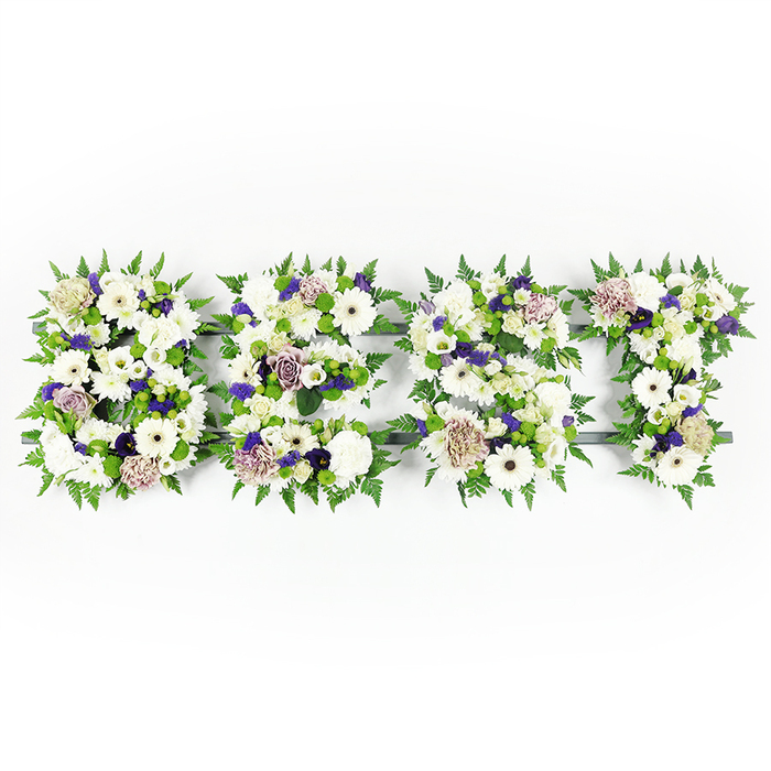 A horizontal, handcrafted floral tribute spells the uppercase word BEST, each letter formed from a rich tapestry of fresh blooms laid out against a bright white surface that makes colours and textures sing. Prominent, pristine white gerbera daisies with their dark centers bring bold, graphic points throughout the letters, balanced by delicate white lisianthus and full, ruffled white carnations that give soft volume and a sensation of tranquility. Soft mauve and lavender roses weave through the composition alongside deeper violet accent flowers, introducing a restrained, respectful warmth to the predominantly white scheme. Feathery green fern fronds trace the outer edges of each letter, their airy fronds creating a natural, scalloped border that both contains and lifts the flower heads. Green button chrysanthemums and compact foliage clusters fill the interiors, adding a fresh, slightly spongy texture that contrasts with the smoother rose petals. The letter forms are anchored on a discreet dark grey base which peeks through in areas, providing structural shape without detracting from the floral surface. The lighting is gentle and even-as you might see in a Clerkenwell studio near Exmouth Market-so whites remain luminous and the mauve tones retain depth. This arrangement reads as a considered sympathy tribute: the whites signify peace and remembrance, the purples a note of reverence, and the overall craft suggests a steady, loving hand. Suitable for wakes or memorial services across Clerkenwell and nearby Farringdon, the piece conveys a message of honour and lasting affection through floral artistry and quiet emotion.