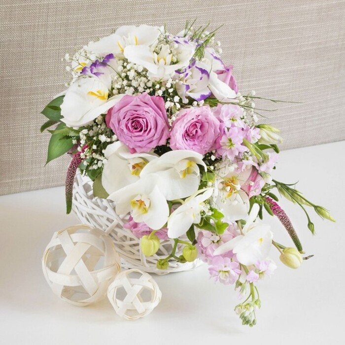 This image shows a luxurious floral table arrangement that looks perfectly at home in a chic Clerkenwell restaurant or wedding venue, captured in a bright, close-up composition. The flowers are gathered in a round white wicker basket, its handwoven pattern painted in a clean, matte white that contrasts subtly with the intricate shadows in the weave. Rising from this basket, a carefully curated selection of blooms creates a gentle dome that slightly spills over the edges, as though the flowers are just on the verge of overflowing. The stars of the arrangement are fully opened pale pink roses, their velvety petals curling outward in classic spirals that suggest softness, devotion, and timeless romance. Interlaced among them are striking white Phalaenopsis orchids, each bloom with broad, satin-like petals and a central column washed with warm yellow and the faintest blush of peach, lending an air of luxury. Surrounding these focal flowers, delicate white Lisianthus with lightly ruffled petals show occasional strokes of lavender along their edges, giving the eye subtle shifts in colour. Wispy clusters of white baby's breath appear between the larger blossoms, adding a fine, airy texture that lightens the composition. Pale pink stock stems trail lightly from the sides, their clustered flowers creating both visual movement and the impression of a soft, sweet scent. Pockets of rich green foliage and a few emerging buds peek through, grounding the pastel tones with a natural vibrancy. The basket sits on a spotless white surface, accompanied by two decorative woven spheres in natural, straw-like material, one large and one smaller, whose open lattice design mirrors the basket's rustic charm. Behind, a neutral beige fabric backdrop with a gentle, linen-like texture frames the scene, ensuring every petal and leaf stands out. The entire arrangement feels calm yet celebratory, ideal for a wedding breakfast near Farringdon or a sophisticated anniversary dinner at home.