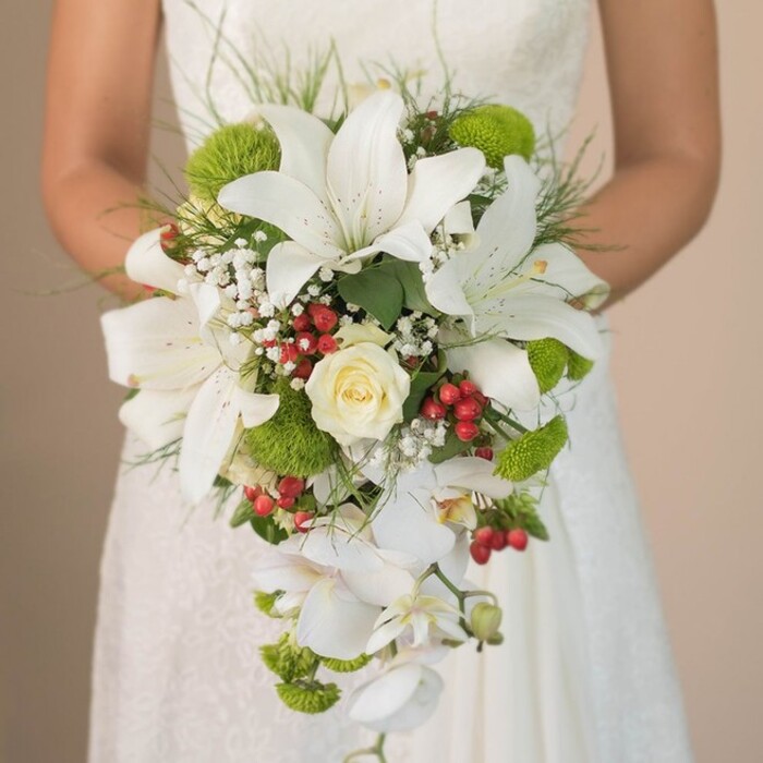 Captured at chest level against a softly blurred backdrop of lace and ivory, a bride in Clerkenwell clasps a luxurious, cascading bouquet bursting with the area's signature creativity and refinement. The striking design features showy, snowy white lilies-each blossom trumpet-shaped and delicately speckled on the inside-forming the sculptural peak of the arrangement. At its heart, a perfectly coiled cream rose suggests classic romance, surrounded by intricate sprays of cloudlike gypsophila, their minute white flowers lending a dreamy softness evocative of morning mist in Spa Fields Park. Contrasting the pale blooms, clusters of lime-green chrysanthemums and pom-like foliage contribute a zesty, contemporary element, reminiscent of Clerkenwell's lively markets and green spaces. Festive red hypericum berries, tucked artfully throughout, add visual warmth and a touch of local festivity, recalling seasonal celebrations along Farringdon Road. Flowing with gentle elegance, multiple stems of trailing white phalaenopsis orchids create a waterfall effect, enhancing the bouquet's sense of movement and luxury. Linear blades of deep green foliage frame the flowers, while hints of rhythmic, grass-like wisps provide structural balance and a natural finish. Sunlight illuminates each petal's satiny texture and nuanced colour, creating depth and dimension. The bride's subtly veiled hands, adorned in lace, cradle the bouquet as though cherishing a work of art. This arrangement-blending tradition with a fresh local vibrancy-stands as an inspirational choice for couples hosting weddings in Clerkenwell's dynamic heart, its lush beauty echoing the spirit of the neighbourhood's elegant celebrations.