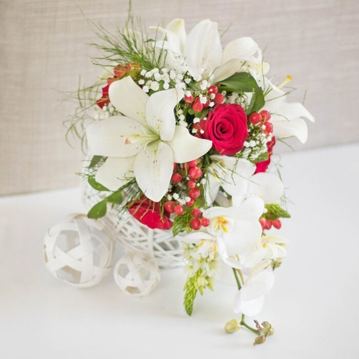 In this image, a refined white and red floral arrangement is carefully styled as if ready to take its place on a wedding table in a Clerkenwell townhouse or chic restaurant near Farringdon. The main bouquet is held in a round, white, open-weave container that resembles hand-plaited wicker, its airy lattice allowing glimpses of stems and giving a light, artisan character to the design. Emerging from this base, pristine white oriental lilies open in generous, star-shaped flowers, each one with smooth, slightly curved petals and golden stamens that stand out against their luminous white. Threaded between these lilies are deeply coloured red roses, their velvety petals tightly rolled in classic spirals, bringing a strong romantic note and a sense of celebration. The larger blooms are softened by sprigs of white baby's breath, creating tiny clusters that look almost like a fine mist of flowers suspended in the air. Bright red hypericum berries and slender sprays of fresh, bright green foliage add an extra layer of festive detail and texture, keeping the arrangement lively rather than formal or stiff. To one side, a trail of white phalaenopsis orchids spills gently downward from the main display, their elegant arch and softly rounded petals giving the piece a sense of movement and grace, as though it were flowing towards the edge of the table. Each orchid bloom has a hint of yellow and soft pink at its centre, barely visible yet adding sophistication. Resting on the smooth white surface to the left are three white woven spheres, echoing the container's pattern and creating a balanced, cohesive tableau. The neutral, light beige fabric backdrop recedes quietly, ensuring the flowers remain the star, ideal for illustrating luxurious wedding décor or intimate celebrations in and around Clerkenwell.