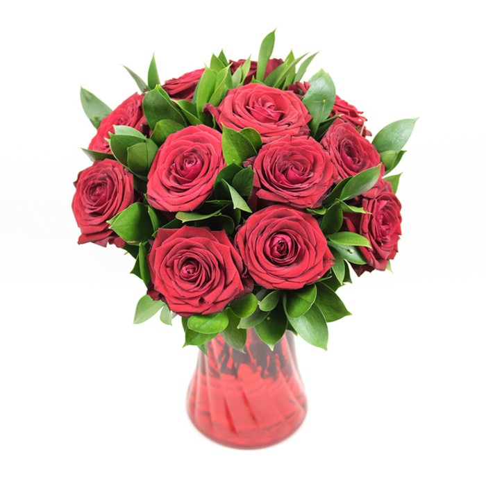 A rich, opulent bouquet of deep red roses is the sole focus of this image, placed in a translucent red glass vase and photographed against a completely white background, as though it were standing on a sunlit windowsill in a Clerkenwell flat overlooking Spa Fields. The arrangement consists of around a dozen to fifteen roses, carefully clustered into a neat, rounded shape where each bloom supports and frames the next. Every rose is open wide, revealing a tightly layered spiral of velvety petals that curl slightly at the edges, suggesting freshness and careful handling by an experienced florist. The red hue is intense and uniform, leaning towards a classic crimson that immediately evokes passion and romance. Tucked generously between the blooms is a halo of glossy dark green foliage, likely ruscus or a similar leaf, adding structural support and a lush, almost woodland feel. The leaves are elongated and slightly pointed, with a smooth, shiny surface that reflects specks of light, creating a dynamic contrast against the matte softness of the rose petals. The glass vase holding the stems is a coordinating shade of ruby red, its body gently curving and narrowing toward the base before flaring outward at the opening. Subtle ribbing or swirling in the glass adds texture and catches reflections, giving the container an elegant yet understated decorative quality. The simplicity of the pure white background isolates the bouquet, amplifying its colours and making it easy to imagine this design arriving by same-day delivery from a local Clerkenwell florist as a romantic gesture for Valentine's Day, an anniversary, or a significant celebration of love.