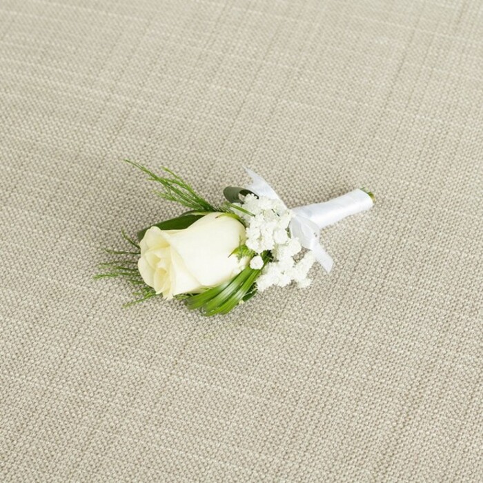 In this carefully composed photograph, a single white rose boutonnière is positioned at a graceful diagonal across a light beige fabric that resembles natural linen, complete with a visible crosshatch texture that feels both refined and organic. The boutonnière's focal point is a creamy white rosebud, its smooth, velvety petals closely wrapped yet softly rounded, as if chosen at the perfect moment before fully opening. Around the base of the rose, hints of green sepals and a ring of slender, deep green leaves create a supportive collar, framing the bloom and drawing the eye inward. To one side, and slightly spilling beyond the line of the rose, sits a delicate cluster of white baby's breath, each tiny blossom forming part of an airy constellation of flowers that lend the design a cloud-soft, romantic quality. Among the foliage, a few finer, fern-like sprigs add complexity and texture, suggestive of natural hedgerow greenery, the sort you might see in pockets of planting near Exmouth Market or along quieter Clerkenwell side streets. The stems of the rose and accompanying foliage are bound together with immaculate precision using a pure white satin ribbon, wrapped snugly to create a smooth, cylindrical handle. Just below the rose, the ribbon is finished with a petite, perfectly symmetrical bow, its soft sheen catching the gentle, diffused light. This finishing touch signals the piece's readiness to be pinned to a suit jacket or waistcoat, perhaps for a ceremony in a converted warehouse venue or a chic city registry office nearby. The overall palette is restricted to calm whites, fresh greens, and the warm neutral of the background, resulting in a timeless, elegant feel-ideal for representing wedding flowers, groom's accessories, and refined event styling in the Clerkenwell area.