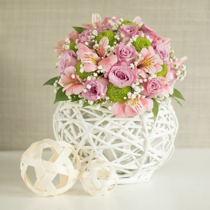 A refined floral arrangement stands confidently on a bright white table, its rounded, abundant form offset by a softly textured beige wall that suggests the calm of an early afternoon in Clerkenwell. The bouquet is built around generous pastel pink and lavender roses, each one fully open so the layered, velvety petals form lush rosettes that seem almost touchably soft. Interspersed among them are graceful stems of pink alstroemeria, their trumpet-shaped blooms marked with fine, painterly stripes of darker pink and warm hints of yellow at their centers, lending delicate intricacy to the composition. Punctuating the pastels are vivid lime green button chrysanthemums, compact and spherical like tiny cushions, adding a fresh, modern pop of colour that energises the palette. Threaded through the flowers are airy clusters of white baby's breath, their tiny blossoms forming a light mist that softens transitions between blooms and enhances the romantic, ethereal mood. Deep green foliage peeks out here and there, providing structure and contrast, like glimpses of a well-tended garden tucked behind the roses. The entire display is cradled in a striking white vase with an open, lattice-like weave, crafted from slender intertwined strips that create a sculptural, basket-style silhouette; its broad, rounded base offers a sturdy anchor for the lavish bouquet. To the left of the vase sit two decorative spheres in pale, natural tones, also woven and openwork, one larger and one smaller, echoing the vase's texture and completing the scene with a sense of balance and thoughtful design. The gentle, even lighting and harmonious colours make this image ideal for imagining a wedding table near Farringdon, a stylish Clerkenwell loft dinner, or a sophisticated gift delivered across EC1 by a florist who understands both freshness and contemporary style.