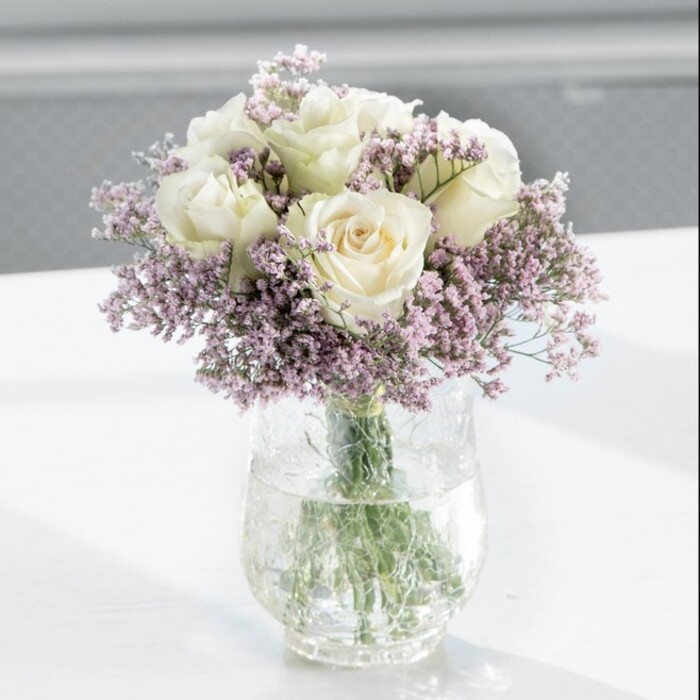 In this image, a gracefully composed, full-bodied bouquet of creamy white roses and lavender statice is the clear focal point, echoing the understated charm you might find in a Clerkenwell gallery space. The arrangement is round and symmetrical, with the roses placed so closely together that they create a smooth, continuous canopy of petals. Each rose appears freshly opened, the outer petals just beginning to curl away from the centre, revealing intricate layers within. Their colour is a soft cream that leans towards ivory, with subtle shading that catches the light and gives the blooms a gentle, luminous depth. Interspersed among the roses, slender stems of statice carry cascades of tiny lilac flowers, adding dimension and texture. These small blossoms have a slightly crinkled, papery feel, contrasting with the roses' velvety surfaces and lending the bouquet an airy, almost weightless halo. All the flowers are gathered neatly into a tall, clear cylindrical glass vase, where the straight green stems are visible in calm, clear water, some angling gently, like lines in a botanical sketch. The vase rests on a smooth white tabletop that reflects hints of green and cream, subtly mirroring the bouquet above. In the background, the scene fades into a soft blur, suggesting an indoor room with daylight streaming in-perhaps similar to an office overlooking Farringdon Road or a peaceful home near Clerkenwell Green. The natural light is bright but diffused, wrapping each petal in a soft glow and casting only the faintest shadows. The overall feeling is one of quiet, modern elegance-an arrangement that could serve as a dignified sympathy tribute, a refined wedding table piece, or a simple, heartfelt gift to bring gentle comfort and poise to any space in EC1.