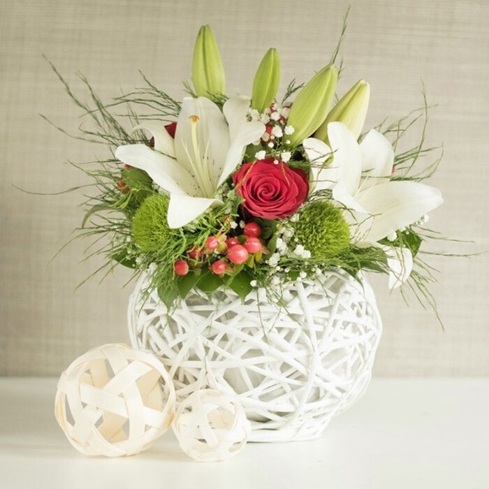 A sophisticated floral display rests on a bright white surface, the scene bathed in soft, even light that would feel at home in a stylish Clerkenwell studio. The focal point is a round, white vase crafted from interlaced strands, its open-weave structure suggesting woven wicker or sculpted branches, giving the piece a handcrafted, contemporary character. From this textured vessel spills a full, thoughtfully layered wedding-style arrangement. Several white lilies stand proudly at the top and centre, some blossoms fully open to reveal wide, gently ruffled petals surrounding golden stamens, while others remain in smooth, elongated green buds that signal ongoing freshness throughout a celebration. Interwoven among the lilies are deep red roses, one particularly prominent bloom near the centre, its tightly curled petals forming a perfect spiral that naturally draws the eye and introduces a note of classic romance ideal for wedding tables in venues tucked around Charterhouse Square or Farringdon. Round, bright green pom-pom flowers-likely dianthus or chrysanthemum-punctuate the design, adding an unexpected, playful texture and vivid colour contrast. Fine sprays of white baby's breath drift like a soft veil between the larger blooms, while clusters of small red berries lend an additional pop of colour and a subtly festive feel. Lush greenery, ranging from delicate, thread-like fronds to sturdier, broader leaves, extends beyond the rim of the vase, creating depth and a sense of natural movement. To the left, two decorative woven balls in lighter natural tones echo the vase's pattern, reinforcing the harmonious, tactile theme. The neutral, lightly textured background ensures that the crisp whites, rich reds, and lively greens remain the undeniable stars, evoking a feeling of refined celebration perfectly suited to wedding receptions and intimate gatherings across Clerkenwell.
