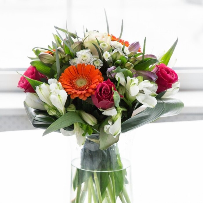 This artful bouquet, showcased against a clean, radiant background, is a graceful expression of both colour harmony and thoughtful design-an embodiment of Clerkenwell's creative spirit. Four prominent gerbera daisies-two ablaze in vibrant orange, two in gentle peach-form a cheerful core, their velvety petals and chocolate-hued centers drawing the gaze with bright, festive energy. Rich fuchsia roses are sculpted into the arrangement with purposeful precision, heightening the sense of romance and luxury. The interlacing curls of white alstroemeria and clusters of tiny white blossoms bring an air of lightness, reminiscent of the tranquil pockets of greenery found near St John's Square. Layered around the base, lush aspidistra leaves are expertly curved by skilled hands, creating a protective frame that enhances both the structure and fullness of the display. Between blooms, pointed green foliage and airy sprigs give the bouquet textural complexity and a gentle, woodland freshness. Transparent glass holds the stems in a natural lattice, reinforcing both the visual and tactile freshness so prized by Clerkenwell Flowers. Sunlight softly illuminates each element, imbuing the colours with vibrancy while preserving a serene balance. Infused with subtle scent and the craftsmanship of a local artisan, this arrangement evokes celebration and thoughtful connection-suitable for gathering with friends on a Clerkenwell terrace or sending as a sign of encouragement to someone embarking on a new journey nearby.