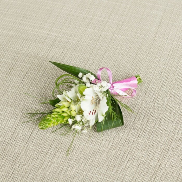A compact, elegant boutonniere is photographed from above, resting on a softly textured, light beige surface that looks like fine linen, reminiscent of table settings in Clerkenwell's boutique venues. The understated neutral background casts no distractions, allowing the floral work to become the gentle focal point. At the centre sits a single white alstroemeria, sometimes called a Peruvian lily, fully open and serene. Its petals are broad and slightly undulating, a creamy white that deepens toward the throat, where slender reddish-brown speckles and streaks gather, giving the flower a painterly, almost calligraphic detail. The stamens sit clearly visible in the middle, hinting at the bloom's freshness and subtle, green-tinged scent. Just to the left rises a tidy, conical cluster of lime-green buds, likely Ornithogalum or a similar spiked flower, each tiny segment closely layered to create a textured, sculptural counterpoint to the softness of the lily. Threaded delicately between these main features are fine stems of white baby's breath, their minuscule blossoms forming airy, cloud-like clusters that lend a romantic haze. The arrangement is anchored by lush foliage: glossy, dark green, sword-shaped leaves offer structure and depth, while slender, grass-like strands of lighter green arc gently outward, adding a sense of movement, as though a breeze from nearby Spa Fields had just brushed across the piece. On the right-hand side, the stems are neatly bound with a narrow, bright pink satin ribbon, tied into a crisp little bow that introduces a playful bolt of colour, suggesting celebration and joy. The lighting is soft and even, enhancing the texture of the petals, the shine of the leaves, and the satin sheen of the ribbon without casting harsh shadows. The overall impression is of a carefully crafted floral accessory-perfect for a groom's lapel, prom night, or any special occasion in and around Clerkenwell-that balances classic white and green tones with a single, cheerful pink accent.