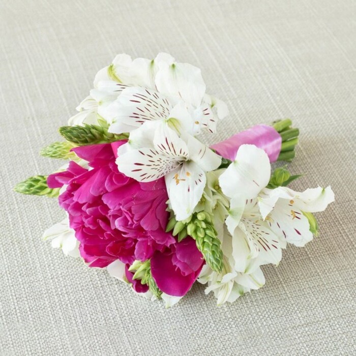 A meticulously designed, compact floral arrangement is laid horizontally on a softly textured, oatmeal-beige fabric backdrop, similar to natural linen used in Clerkenwell studio spaces. The eye is immediately drawn to a single, lush magenta bloom at the centre-its shape and petal structure recalling a small peony or heavily ruffled garden rose. Countless petals overlap in a tight, domed formation, each edge slightly frilled, creating a sense of plush depth and softness, as if the flower were a cushion of rich velvet. Encircling this bold focal blossom are several white alstroemeria, or Peruvian lilies, their trumpeted flowers opening with quiet grace. Each petal is long and slender, slightly curling back to reveal intricate inner markings: fine reddish-brown and burgundy streaks and freckles that fan out from the throat, suggesting both delicacy and wildness in a single stroke. Threaded among these blooms are narrow, light green buds and sprigs of fresh foliage, some upright and spiky, others gently arching, adding structure and an airy, garden-like freshness. The stems have been precisely gathered and wrapped in a glossy satin ribbon dyed to a bright fuchsia that echoes the central flower's hue, the ribbon smooth and neatly finished, giving the design a polished, couture feel. Soft, even lighting captures the interplay of textures-the matte petals, satin sheen, and gentle weave of the fabric-while the neutral background keeps the focus entirely on the colour harmony of pink, white, and green. The piece feels perfectly sized for a bridal party accessory, perhaps a boutonnière or wrist corsage for a wedding at a Clerkenwell church or a chic celebration in a nearby warehouse venue, embodying modern London romance with a refined yet joyful character.