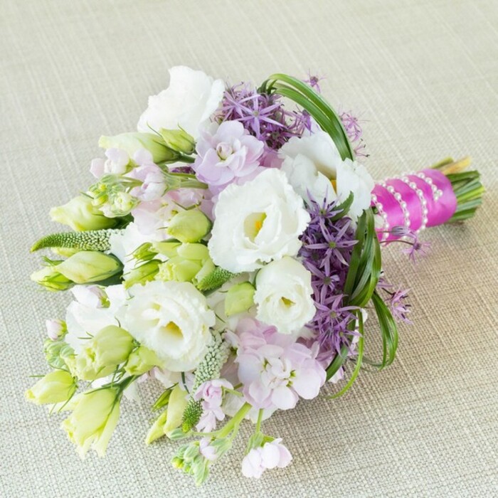 Set against the understated elegance of a lightly textured cream linen-a nod to Clerkenwell's blend of historic charm and modern design-this refined floral bouquet overflows with thoughtful detail and local character. Meticulously arranged, the bouquet's focal point is a sweep of full, ruffled white Lisianthus (Eustoma), their petals luminous under the soft, even lighting, and their centres kissed with pale yellow. Wisps of white and lime-green lisianthus buds emerge throughout, imbuing the composition with freshness, while clusters of pastel lavender-pink stock offer a fragrant and gentle counterpoint. Providing dynamic contrast, deep purple allium flowers with their characteristic starburst florets introduce unexpected architectural flair, reminiscent of public art and creative spaces that define the area. Weaving through the bouquet, slender green bear grass and textured fillers bring texture and movement evocative of Clerkenwell's hidden gardens and tree-lined boulevards. The stems are expertly gathered and finished with a vivid fuchsia satin ribbon, neatly intertwined with a strand of glossy white pearls for a bridal touch brimming with style and tradition. This bouquet is perfectly suited for modern weddings or urban celebrations in Clerkenwell, a floral arrangement that echoes the neighbourhood's harmony of the contemporary with the classic, embodying themes of love, renewal, and understated luxury.