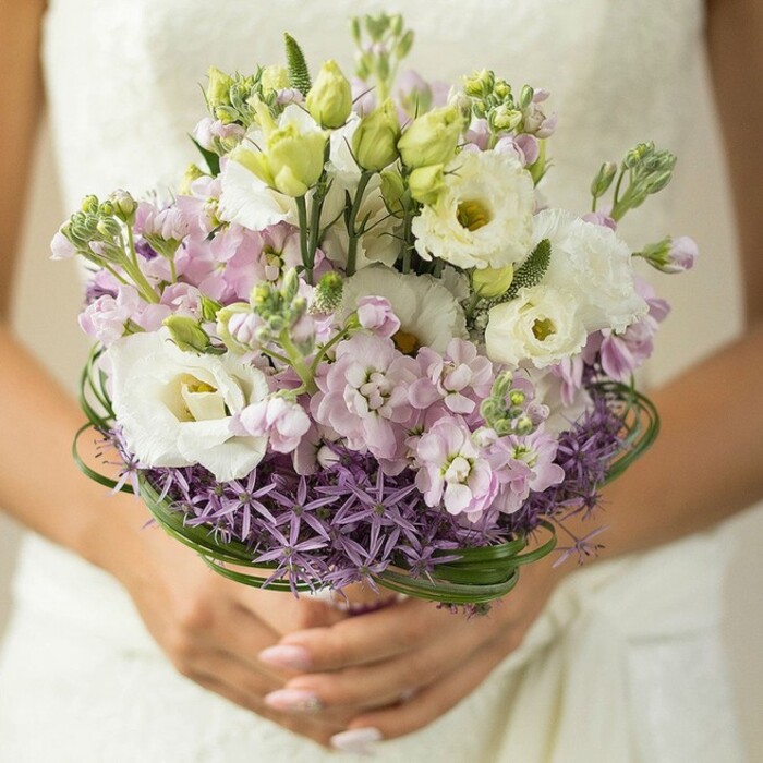 In a luminous, softly-focused close-up, a bride holds an exquisitely crafted bouquet-a work of floral artistry echoing the elegance of a Clerkenwell wedding. Dominating the composition are sumptuous white lisianthus blossoms, their petals layered in gentle ruffles, attended by clusters of pale pink and lilac stock whose petite, fragrant blooms create a dreamy, textured canopy. Slender buds and fresh green accents mingle amongst the pastels, while a band of richly colored purple allium traces the bouquet's base, lending sculptural contrast and a touch of whimsy. Above the allium, gracefully looping bear grass provides both structure and a nod to contemporary design, all meticulously assembled by the local Clerkenwell Flowers team. The bride's hands, elegantly manicured with a subtle nude sheen, cradle the bouquet above an ivory lace dress, softly blurred in the background, evoking a sense of anticipation before a ceremony at St. John's Gate. This lush, ethereal bouquet balances tradition and modernity, reflecting Clerkenwell's unique blend of history and artistry as it embodies love, excellence, and the promise of new beginnings.