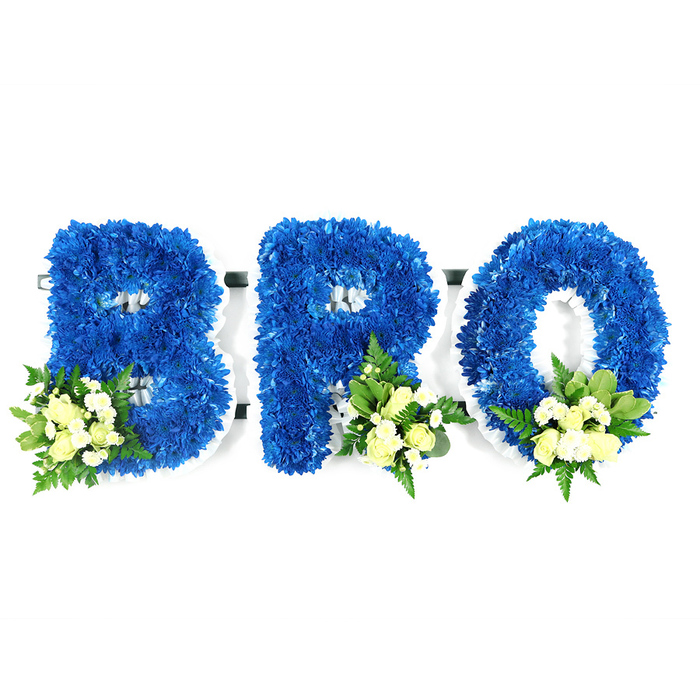 Framed against an immaculate white background, this three-part memorial spells BRO in three substantial capital letters formed entirely from saturated royal blue chrysanthemums. The design is compact and deliberate: thousands of small florets are tightly packed to produce a lush, tactile face on each letter so they read as solid, comforting shapes. At the lower left of the B and at the lower right of the R and O sit small bouquets of soft, pale yellow to ivory spray roses, paired with tiny white filler blooms - perhaps mini chrysanthemums or delicate gypsophila - and layered with deep glossy salal leaves and airy, fern-like greenery. Those accent clusters break the intensity of blue with a gentle, candlelit glow and add both visual focus and soothing texture.
A slim white edge and interior base are evident where the letters' counters open, suggesting the supportive foam or frame beneath the flowers and communicating the craftsmanship involved in building a freestanding tribute. The lighting is soft and neutral, chosen to reveal petal texture and the subtle interplay of colour without casting heavy shadows. The overall impression is reverent and composed: blue for fidelity and calm, cream for solace and purity. This kind of personalised tribute is often selected by families across Clerkenwell, around Charterhouse Square and the nearby Hatton Garden area when they need a dignified expression of condolence for a brother or close friend.