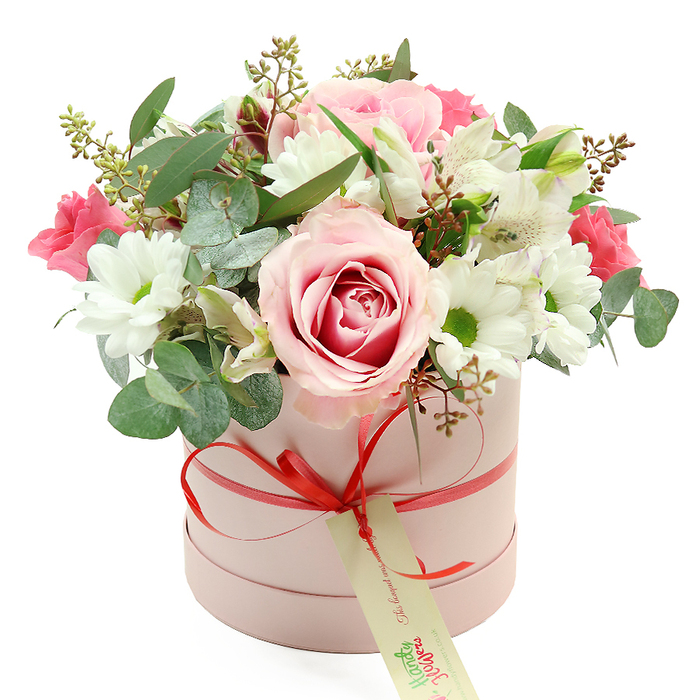 Against a bright white background, a round blush-pink hatbox stands proudly, cradling a lush bouquet that feels both contemporary and timeless, much like the blend of old and new architecture around Clerkenwell. The box has a soft, matte surface in a gentle pastel pink, its cylindrical shape echoing traditional hatboxes but with a modern, minimalist twist. Just above the base, a slim coral-red satin ribbon encircles the box and is tied into a small, perfectly formed bow at the front. Suspended from the bow is a cream-coloured gift tag with green, handwritten-style text, suggesting a personalised note from someone who has chosen their flowers with care. From the open top of the box, the flowers surge outward in a rounded, domed canopy of petals and leaves. At the forefront sits a large bi-colour rose, its outer petals pale blush and its centre swirling into a richer pink, fully unfurled and plush. Nestled nearby are clusters of pure white chrysanthemums or daisy-like blooms, each with a vivid yellow-green centre that brings brightness and clarity to the design. Among them, white alstroemeria stretch out slender, trumpet-like petals marked with delicate speckles, lending an airy, graceful quality. Intermixed are smaller, vibrant deep pink flowers, such as spray roses or lisianthus, that punch through the softer tones with little bursts of fuchsia. The bouquet is framed and filled with an abundance of greenery: rounded, silver-green eucalyptus leaves that arc gently and suggest a fresh, clean scent; deeper green leaves that provide contrast and depth; and fine stems tipped with tiny green buds or muted brown seed heads for natural texture. The result is a thoughtfully crafted floral hatbox, ideal for local delivery to a townhouse near St John Street or a design studio by Clerkenwell Green, perfect for birthdays, anniversaries, or simply bringing a sense of gentle celebration into a room.