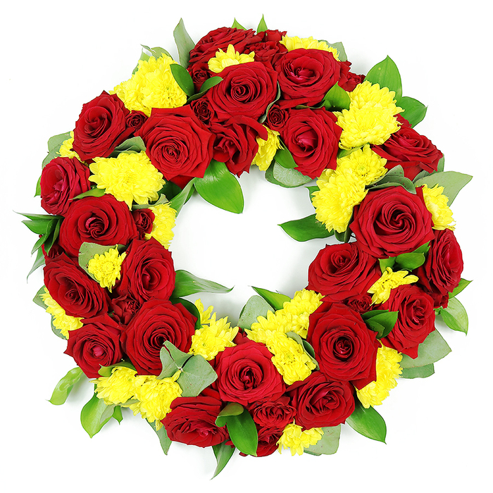 This image shows a beautifully composed circular funeral wreath, photographed against a crisp white backdrop so that every flower stands out clearly, as if displayed on a stand in a quiet Clerkenwell chapel. The wreath is lavishly packed, forming a complete and unbroken ring symbolising continuity, remembrance, and enduring affection. Dominating the design are numerous red roses at varying stages of full bloom, their petals rich and velvety, layered in tight spirals that open outwards into soft curves. The shades of red move gently from bright crimson on the outer petals to deeper, almost wine-like tones closer to the centre, creating a sense of depth that feels both romantic and reverent. Threaded generously among these roses are groups of small yellow chrysanthemums, their many fine petals forming rounded, button-like blooms that add texture and cheerful warmth to the arrangement. The yellow appears bright yet gentle, like a ray of sunshine on a sombre day, bringing a quiet sense of hope to the tribute. Beneath and around the flowers, an abundance of foliage creates a lush green framework: broad, healthy leaves with a subtle gloss, likely salal or similar, are interspersed with sprigs of softer, grey-green leaves that resemble eucalyptus, adding a slightly misty, calming note. The foliage is neatly tucked so that it supports rather than overwhelms the blossoms, ensuring the red and yellow palette remains the focal point. The even lighting reveals each detail-the crisp edges of petals, the layered greens, the clean inner circle of the wreath-making it an ideal visual representation of a respectful sympathy tribute for services and memorials across Clerkenwell and the surrounding streets.