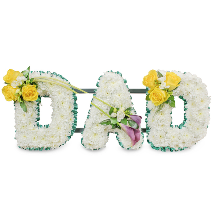 A carefully composed memorial spelling out DAD in large, freestanding letters, each letter cloaked in a dense carpet of immaculate white blooms - small spray chrysanthemums or spray carnations - which give the letters a rounded, plush quality. The perimeter of every letter is traced with a ruffled, dark-green satin ribbon that crisply defines the silhouette and provides a vivid contrast to the white flowers. Behind the letters, slim dark-green metal bars are visible running horizontally, the subtle structural frame that keeps the trio aligned and upright. On the left 'D', a luminous cluster of bright yellow roses nestles into the upper left curve, paired with tiny white freesias and fresh green foliage; from this grouping two long, graceful bare green stems extend diagonally toward the central letter, creating a gentle sweep across the arrangement. The central 'A' carries a modest yet striking embellishment near its lower left diagonal: two deep purple to burgundy calla lilies whose smooth, sculpted forms command quiet attention, set amidst small white filler blooms and verdant leaves. The right 'D' mirrors the left with the same cheerful yellow rose cluster on its upper right curve, producing a harmonious visual echo. Set against a clean white background in soft studio light, the combination of textures - velvety calla petals, crisp ribbon, plush white florets and glossy foliage - reads as dignified and comforting. The overall impression is one of respectful remembrance: a bespoke funeral tribute that visually speaks of love, friendship and reverence, suited to memorials in Clerkenwell and nearby meeting places such as St John's Square and the markets around Smithfield.