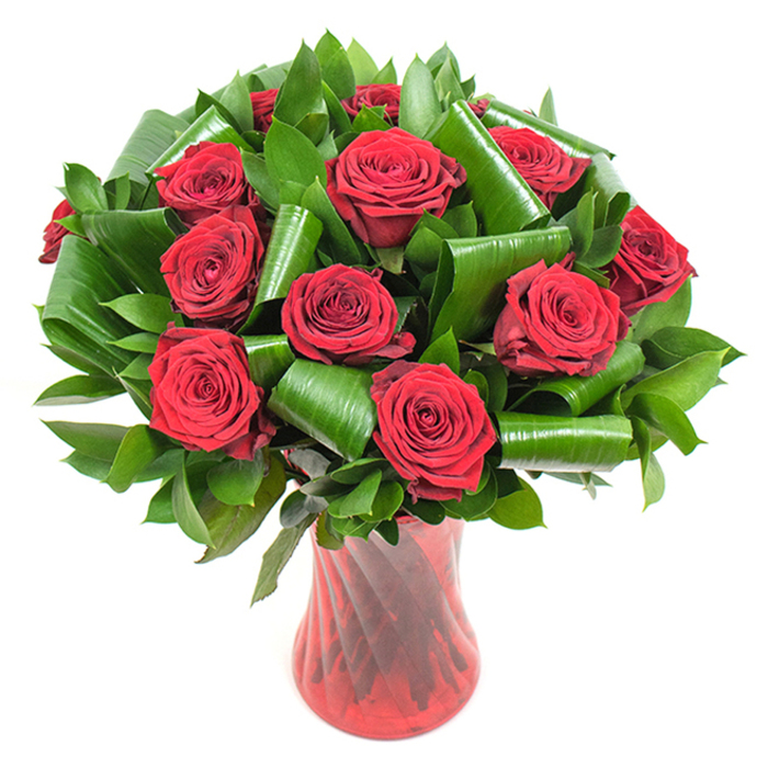 This carefully composed arrangement presents approximately a dozen deep crimson roses, each bloom opened to reveal tightly spiraled, velvety petals that absorb and reflect light like plush fabric. The roses form a lush, rounded mass, their rich red tones intensified by soft, even illumination that brings out petal texture and subtle tonal variation from core to edge. Surrounding the blooms is an abundance of dark green foliage: smaller, glossy leaves create a verdant cushion while broader leaves - reminiscent of Aspidistra - are expertly rolled and curved into sculptural cylinders and loops. Those shaped leaves are not merely filler but structural elements, artfully placed to cradle stems, add height, and introduce elegant architectural movement that modernises the classic red-rose silhouette.  A tall, translucent ruby-red glass vase with a gentle waist and a faint ribbed twist completes the composition; its vertical lines echo the stems and reflect light in a way that unifies vase and flowers. The whole piece is photographed against a pure white background so the deep reds and emerald greens read with vivid clarity. Crafted in our Clerkenwell studio by experienced hands at Clerkenwell Flowers, this bouquet balances traditional romance with contemporary design - ideal for anniversaries, Valentine's gestures, or refined venue styling near Charterhouse Square or Smithfield. The image invites you to imagine the soft, heady scent of rose and the smooth, cool feel of glass beneath your fingertips, promising a memorable floral moment for someone special.