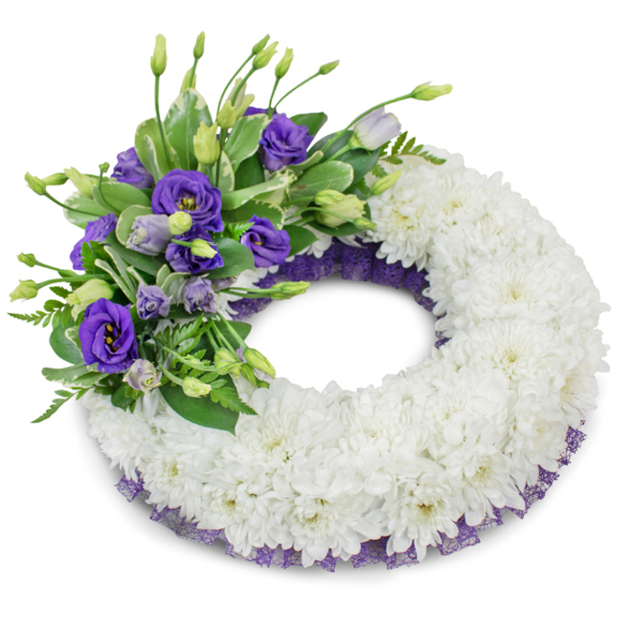 A studio image of a circular funeral wreath, where the visual language is one of soft restraint and considered detail. The foundation is a dense, uniform ring of snowy white cushion chrysanthemums - each bloom's tightly layered petals nestling against its neighbours to create a seamless, tactile surface that suggests comfort and continuity. At the upper-left quadrant a composed accent of purple Eustoma (lisianthus) introduces warmth and colour: richer violet blossoms sit beside paler lavender flowers and slender unopened buds, their layered petals offering a velvety texture against the chrysanthemum's ruffle. Mixed among these purples are trimmed sprigs of green foliage - some glossy and broad with subtle variegation, others fine and fern-like - which provide natural rhythm and a cooling counterpoint to the warm purple. A delicate band of dark purple mesh or lace is visible beneath the white blooms along both inner and outer edges, a restrained decorative touch that harmonises with the lisianthus and gives a refined outline to the wreath. The arrangement is lit softly so that a gentle shadow falls beneath, giving the piece presence and dimensionality on the plain white field. This memorial wreath, with its classic white and symbolic purple palette and carefully balanced textures, is intended for funerals and graveside tributes where quiet dignity and lasting remembrance are paramount.