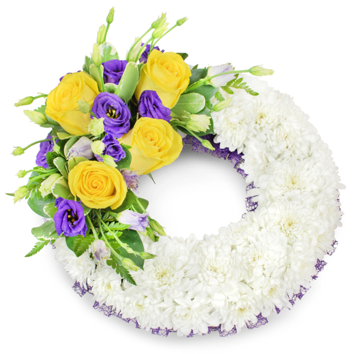 Against a seamless white background, this traditional circular funeral wreath is constructed with the quiet precision of an experienced florist's hand. The dominant surface is a continuous ring of pristine white spray chrysanthemums, packed densely so that their fine, overlapping petals create a cushion-like, clouded texture that reads as purity and collective comfort. A concentrated floral spray sits on the upper left arc: several rich yellow roses, fully unfurled in warm honey and butter tones, offer a soft perfume and a feeling of gentle companionship, while deep purple lisianthus (Eustoma) punctuate the composition with their velvety, ruffled, cup-shaped flowers. A few pale lavender and green lisianthus buds remain closed, introducing dimension and a natural pause. The accent is layered into fresh green foliage - glossy rounded leaves, subtle variegation, and feathery fern fronds - which holds the blooms and gives the cluster its graceful sweep. A slim band of dark purple mesh or crinoline is visible beneath the outer and inner edges of the chrysanthemum ring, a refined echo of the lisianthus hue that frames the piece. The wreath's hollow centre emphasizes the traditional 'O' silhouette and the symbolism of continuity and remembrance. Bathed in soft, neutral light the textures come alive: the chrysanthemums' softness, the roses' buttery density, and the lisianthus' satiny coolness. As a local tribute crafted with care, this wreath is the kind of sympathetic design Clerkenwell Flowers might supply for memorials near Exmouth Market or Smithfield, combining quiet dignity with a gentle, warm accent of colour.