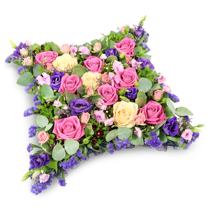 A beautifully detailed funeral cushion is displayed here, shaped like a soft, pillow-like rectangle with rounded edges that evoke rest and serenity. Every inch of the cushion is densely planted with fresh flowers, creating a lush, seamless surface of colour and texture. The main focus is on generous roses in varied tones: bold fuchsia and raspberry pink sit alongside tender blush and pale pastel pinks, balanced by creamy, almost buttery yellow roses. Many of these roses are fully open, revealing layered petals that look almost like finely folded silk. Interspersed among them are velvety, royal purple lisianthus blooms, their deeper shade adding contrast and a quiet, dignified mood. Smaller spray roses in delicate pinks, along with tiny clusters of lavender-blue filler flowers resembling statice or limonium, tuck into the spaces between larger heads, giving the design a rich, intricate pattern. Surrounding and underpinning the blooms is an abundance of greenery, including cool, rounded eucalyptus leaves with their soft silvery-green hue and darker, feather-like foliage that adds structure and depth. The arrangement is photographed against a clear white background, a soft shadow suggesting its weight and three-dimensional form, as though it rests on a workbench in a Clerkenwell flower studio near Spa Fields. This tribute feels thoughtfully composed for a memorial, funeral, or graveside service in the local area, conveying condolences, love, and remembrance in a way that speaks gently but clearly, much like a whispered farewell shared along the quiet streets near St John's Square.