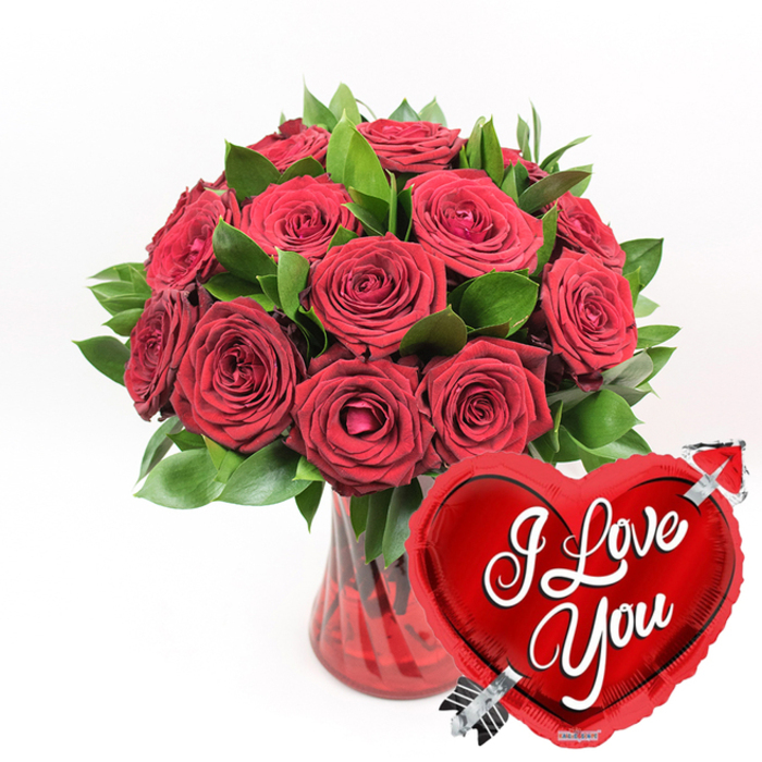 Against a flawless white background, a sumptuous bouquet of deep crimson roses and glossy greenery captures the eye, radiating the kind of polished romance you might associate with a carefully prepared surprise in Clerkenwell. The arrangement contains approximately a dozen or more roses in varying stages of bloom, from tight, promise-filled buds to fully opened blossoms revealing layers of soft, velvety petals. Their colour is a rich, uniform red, the shade that instantly evokes love letters and evening dinners. The roses are set among lush green foliage, leaves clustered densely enough to frame each flower while still letting the blooms remain the centre of attention. They stand in a translucent ruby-red glass vase, its fluted sides shaped into gentle vertical curves that create repeating highlights and shadows as light passes through, adding a subtle sense of movement and refinement. In the lower right area of the composition, slightly overlapping the base of the bouquet, a bright, metallic red heart-shaped foil balloon introduces a playful counterpoint to the classic florals. The balloon's high-shine surface reflects light in silvery sparkles, and across its centre, the words I Love You are written in smooth, white cursive lettering that arcs comfortably within the heart's outline. A silver Cupid's arrow seems to pass through the balloon on a diagonal, suggesting the traditional imagery of love struck at first sight. With no other visual distractions, the clean white setting allows the rich reds and greens to stand out boldly, making this image an ideal representation of a romantic gift ready to be sent to a home or restaurant in Clerkenwell for Valentine's Day, an anniversary, or any heartfelt declaration of affection.