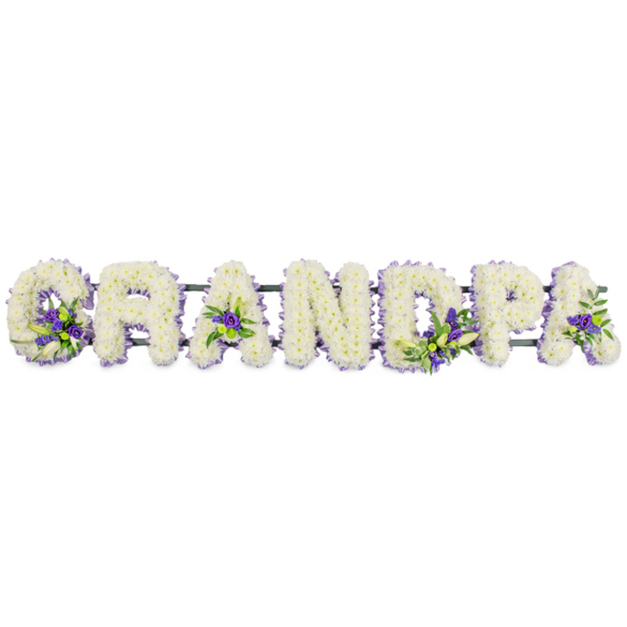 This photograph captures a personalised funeral floral tribute arranged to spell the word GRANDPA in sturdy block capitals, presented in a clear horizontal line against a bright white backdrop so that the design appears almost to float. Every letter is generously covered in small white blooms - most likely chrysanthemums or carnations - arranged so closely together that they create a thick, plush carpet of petals. The white flowers give the letters rounded edges and a soft, cloud-like texture, suggesting calm, purity, and the gentle passing of time. Deliberate accents of deep purple and lavender flowers sit within and around the shapes of the letters, joined by sprigs of fresh green foliage. These splashes of colour nestle in familiar places: near the lower curve of the G, resting in the central triangle of the first A, threading through the D, softening the bowl of the P, and finishing the final A with a touch of warmth. The combination of white and purple feels balanced and thoughtful, reminiscent of the quiet elegance seen in memorials near Clerkenwell Green or along the paths of nearby historic churches. Beneath the blooms, a low, dark green frame runs from one end of the tribute to the other, efficiently supporting the weight of the flowers while remaining almost invisible to the eye. This design would typically be placed alongside a coffin, at the front of a chapel, or by a graveside as family and friends gather, its presence quietly stating the grandfather's role in the family. The arrangement's neat lettering, precise workmanship, and carefully chosen colours make it a touching way to express love, gratitude, and final farewell within a local Clerkenwell funeral service.