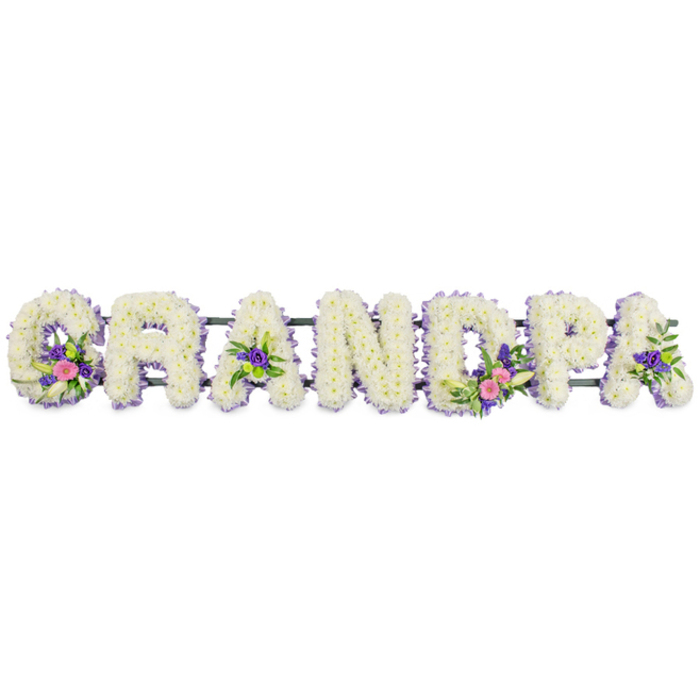 A bespoke floral name tribute spelling GRANDPA is laid out horizontally against a stark white background, its craftsmanship suited to a thoughtful funeral or memorial service in Clerkenwell's historic churches and crematoria. Each bold capital letter is built from a densely packed bed of delicate white flowers, most likely spray chrysanthemums or tiny carnations, their many petals overlapping to form a velvety, cushion-like surface that looks both soft and abundant. Encircling every letter is a fine margin of petite purple and lilac blooms, perhaps statice, heather, or asters, creating a cool lavender outline that crisply defines the shape of each character and gently lifts the design from the background. Within the word, specific letters carry carefully arranged accent sprays that add a sense of individuality and remembrance: the 'G' hosts a cluster of bright pink flowers and a slender white lily-like bloom, complemented by fresh, glossy foliage; the first 'A' includes smaller touches of deep purple; the 'D' repeats the lily and pink theme with extra vibrancy; and another 'A' carries similar purples and greens, including hints of bright green button chrysanthemums. These focal points bring movement and depth, so the tribute feels more like a living memory than a static display, echoing the way families around Clerkenwell tell stories of their grandfathers over walks near Exmouth Market or along Farringdon Road. A slim, dark green horizontal bar runs behind the letters, partially visible between them, acting as a subtle frame and support while ensuring the name reads clearly from any angle. The overall mood of the composition is quiet, dignified, and deeply personal, a floral lettered tribute designed to honour a much-loved grandfather with grace and tenderness.