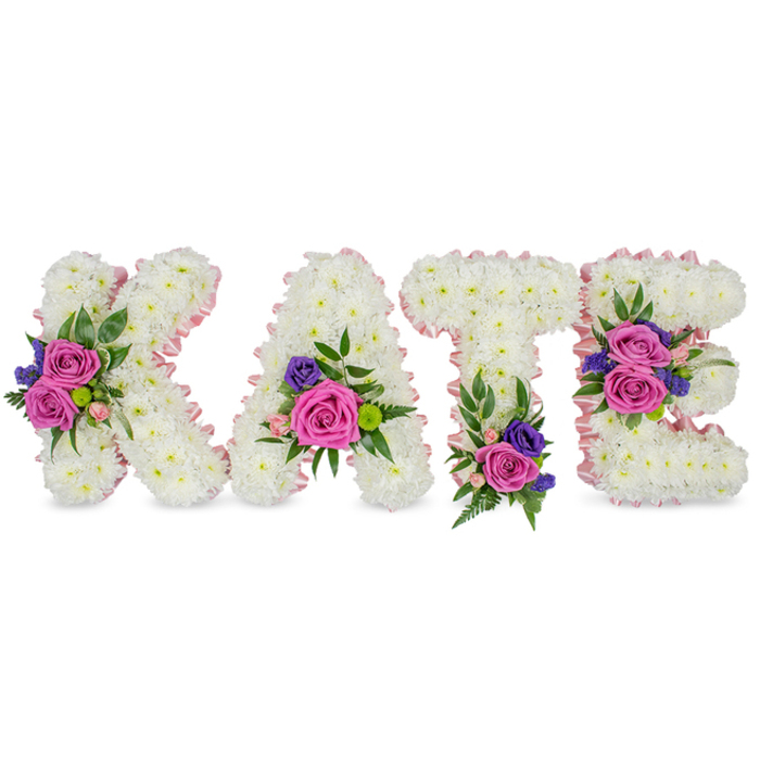 Here a bespoke floral name tribute spells KATE in four upright, freestanding letters formed from thousands of small, immaculate white chrysanthemums that together present a soft, dense texture like a snowy thicket. Each letter is carefully edged with a lustrous pale pink satin ribbon that defines the shapes and adds a whisper of warmth to the stark white field. Clusters of accent flowers interrupt the white with concentrated bursts of colour: bold fuchsia-pink roses paired with delicate pastel pink spray roses and fine stems of deep purple or blue blooms, perhaps statice or small lisianthus, chosen to give depth and a touch of solemn richness. The floral groupings are framed by varied greenery - slender fern fronds, rounded eucalyptus leaves and occasional glossy foliage - which lend contrast, movement and a soothing green undertone. The arrangement's composition is deliberately balanced: the K shows a prominent diagonal cluster, the A carries its posy near the lower right curve, the T is anchored by a grouping at its base, and the E bears two clusters on the middle and bottom bars; together these placements guide the eye gently across the name. The overall impression is one of tender craftsmanship and reverence; the roses' velvety petals and the chrysanthemums' soft heads combine to create a comforting tactile image and a subtle perfume. Designed to honour a life, this type of tribute is often chosen for funeral chapels, memorial services or graveside ceremonies, and would be at home for families in Clerkenwell recalling walks through Leather Lane or quiet moments at nearby Spa Fields.