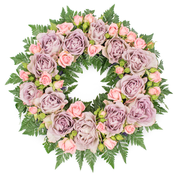 A large, circular funeral wreath is shown laid flat on a pristine white surface, the overhead view allowing every detail of the floral design to be appreciated. The ring is composed of lush roses in a quietly sophisticated colour palette: muted lavender, dusty mauve, and soft grey-purple tones swirl together as each bloom opens fully, revealing finely ruffled petals that layer like silk. Between these statement roses are clusters of petite blush pink spray roses, some still enclosed in tight, hopeful buds and others just beginning to unfurl, creating a gentle rhythm of shapes and sizes that softens the overall look. The flowers are anchored in a generous collar of deep green fern fronds, probably leatherleaf and similar foliage, their serrated, feather-like leaves radiating outwards and creating a textured green halo around the pastel centre. Subtle accents of darker filler greens and tiny, near-black buds are tucked among the roses, adding contrast and a slight shadow that makes the blooms seem to float above the foliage. The composition is perfectly round and carefully balanced, symbolising continuity and eternal love, which makes it especially fitting as a sympathy or memorial tribute. The mood captured in the photo is reflective and calm, like the quiet streets around Clerkenwell's historic squares in the early morning. This is the sort of wreath a local family might choose for a funeral service at a nearby church or crematorium, or to rest beside an urn at a celebration of life, its soft lavender and pink tones sending a gentle message of remembrance, grace, and lasting affection.