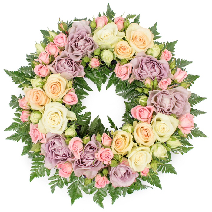 In this image, a full circular wreath of roses and foliage is centred against a pure white background, the simplicity of the setting drawing attention to the careful craftsmanship of the flowers. The wreath is abundant and tightly packed, leaving no gaps, so that the eye travels around an unbroken ring of colour and texture. Dominating the design are large roses in soft, muted shades: creamy white, pale peach, and a distinctive dusty lavender that feels both modern and timeless. Their petals, slightly ruffled at the edges, suggest softness to the touch, as if they would yield under a gentle fingertip. Between these focal blooms, delicate pink spray roses cluster in small groups, mixed with pale green buds that hint at new life and the ongoing cycle of nature. The darker, finely divided fronds of fern and similar greenery sit beneath and between the roses, providing structure and a cool, forest-like backdrop to the paler tones above. The green foliage also traces the inner and outer edges of the circle, accentuating its symbolic shape of eternity and remembrance. The overall palette is calm and restful, suited to a funeral or memorial tribute for a loved one from Clerkenwell, perhaps someone connected to the creative studios around Farringdon or the historic streets near St John's Gate. There is a sense of quiet dignity in the composition, as though each flower has been placed with intention and care. The wreath appears fresh and vibrant, suggesting it has been prepared recently by a local florist who understands how flowers can speak softly when words feel difficult.