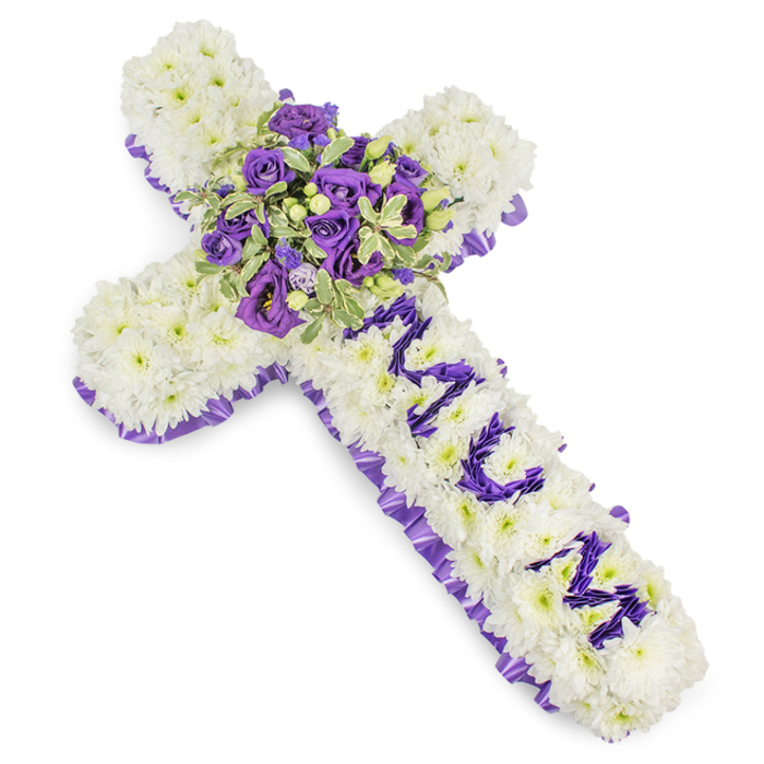 A large, expertly constructed Christian cross of flowers rests against a stark white background so every botanical detail is visible: the main field is a dense layering of small white chrysanthemum blooms, their rounded petals forming a soft, pillowy surface that absorbs light and suggests purity. Around the entire perimeter a deep purple satin ribbon has been artfully pleated and secured, providing a formal, textured frame that heightens the design's solemnity. Running down the vertical arm, the word MUM is spelled out in distinct letters using compact purple flower heads - likely eustoma or fine spray chrysanthemums - the colour chosen to read clearly from a distance and to personalise the tribute. At the cross's centre a generous spray forms the focal point: rich violet roses with tight, plush blooms sit with lighter lavender lisianthus whose ruffled edges create movement; variegated greens, possibly pittosporum or similar foliage with pale-edged leaves, introduce brightness and definition, while little green berries add punctuated texture. The tonal range from pristine white through soft lavender to deep violet creates a palette of reverence, calm, and dignity. The textures - satiny ribbon, velvety rose petals, downy chrysanthemum heads, and crisp variegated leaves - contrast and complement one another, offering both visual depth and a tactile sense of consolation. This arrangement reads as a carefully considered memorial piece, suitable for funeral services in Clerkenwell, Finsbury or at the local church near Clerkenwell Green, crafted to honour and remember with lasting affection.