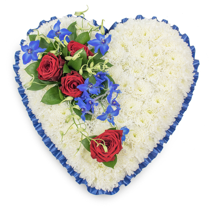Elegant heart-shaped sympathy arrangement designed as a funeral tribute, laid flat and photographed from above so the symmetry and texture are clearly shown. The foundation of the design is a densely packed base of white chrysanthemums, their short stems and rounded heads forming a uniform carpet of petals that looks peaceful and almost cloud-like. Around the outer contour of the heart runs a wide royal blue satin ribbon, precisely pleated or ruffled so that each fold stands out, giving the piece a polished, ceremonial frame. In the upper left portion of the heart, a carefully composed spray draws the eye: three fully open, deep red roses, velvety and rich, nestle among dark glossy leaves that cradle and support them. Threaded through this cluster are bright blue dendrobium orchids, their star-shaped blooms arching and spilling softly across the mums, with fine, curling green tendrils adding a touch of lightness and organic movement. On the lower right curve, a solitary red rose set into the white base offers a quiet echo of the main spray, like a single voice of remembrance answering the group above. The combination of white, red and blue suggests purity, love, loyalty and calm, creating a mood that is both strong and soothing. It feels like a tribute that could rest gently in a Clerkenwell chapel or be carried through the narrow streets near Farringdon as part of a farewell service. Every detail-from the crisp mums to the satin ribbon edge-reflects the care and respect behind a thoughtfully arranged memorial heart, intended to honour a life with grace and beauty.