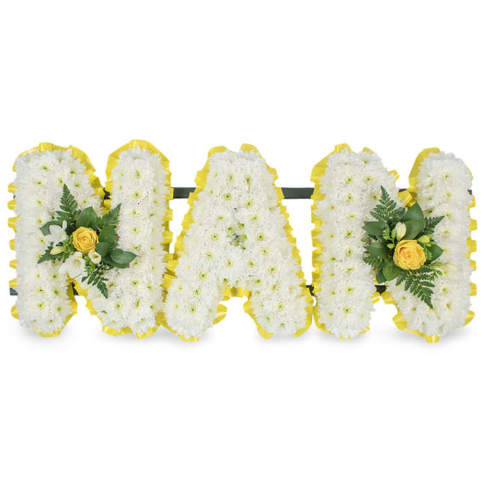 A bespoke MAM tribute arranged with loving precision, each capital letter densely packed with small, pure white chrysanthemums to create a continuous, tactile surface that reads as both delicate and substantial. The full perimeter of each letter is trimmed with a bright, cheerful yellow satin ribbon, pleated for texture and laid with exacting care so that the sunny band outlines every curve and angle, giving the composition a crisp finished edge against a pristine white background. The letters are mounted on a slim, dark horizontal support that sits behind their upper sections, indicating a freestanding piece designed to be placed at a funeral service, memorial or graveside. Two identical floral embellishments accent the arrangement: one is set on the left upright stroke of the first M and the other mirrors it on the right upright stroke of the final M. Each cluster centres on a softly furled yellow rose bud, encircled by sprays of tiny white freesia or similar buds and layered with dark green fern fronds and glossy foliage that add depth and a natural contrast to the whites and yellows. The photograph's soft lighting brings out the satiny sheen of the ribbon, the fine texture of chrysanthemum florets, and the lush, cool greens of the accompanying leaves. As a local Clerkenwell florist might note, this composition balances reverent simplicity with a warm accent-an expressive tribute suited to services near St John's Square, at Farringdon chapels, or for a family laying a mother to rest.