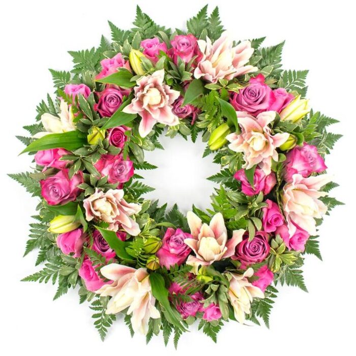 A detailed image shows a full, circular funeral wreath laid out against a spotless white background, each flower and leaf standing out with almost painterly clarity. The design is dominated by roses in a rich spectrum of pink tones, from intense magenta and fuchsia to softer, romantic blush. Some roses are at their peak, fully open with layered, velvety petals curling gently at the edges, while others sit as neat buds or half-open blooms, suggesting the passage of time and the many chapters of a cherished life. Threaded between the roses are generous heads of double lilies, possibly oriental hybrids, whose petals overlap in multiple tiers of creamy white, pale pink, and hints of peach, with occasional darker pink veins tracing toward the tips. A handful of bright, lime-green lily buds rise from the design, bringing a fresh, almost springlike energy. The entire ring is supported and framed by a deep bed of green foliage: finely cut fern fronds creating a soft, airy fringe, and glossier, broader leaves adding contrast and depth, much like the mix of wild greenery and cultivated planting you might notice walking through Clerkenwell's hidden gardens and courtyards. The wreath forms a smooth, unbroken circle with no gaps, conveying completeness, unity, and eternal remembrance. The stark white background allows the rich colours to glow, heightening the sense of care, craftsmanship, and dignity. This arrangement would feel perfectly at home as a tribute at a Clerkenwell funeral service, memorial gathering, or celebration of life, offering a gentle yet striking expression of sympathy and enduring love.