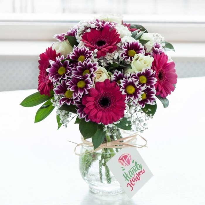 A richly coloured, mixed flower bouquet is captured in crisp detail, poised in a low, clear glass jar-style vase against a bright, softly blurred white background that evokes the calm light of a Clerkenwell apartment near Farringdon station. The central stars are large gerbera daisies in a vivid fuchsia or magenta, their smooth petals forming perfect circles around deep, dark centres that add dramatic contrast. Surrounding them is an artful ring of daisy-like chrysanthemums, each bloom painted with purple at the outer tips of the petals, fading inward to pure white, and finished with a bright yellow centre that glows warmly under the natural light. Interspersed throughout the arrangement, creamy white spray roses open in gentle spirals, with some buds still closed, promising more beauty to come and lending an air of romance and refinement. Fine sprays of white gypsophila thread delicately between the larger blooms, their tiny, cloud-like flowers giving the composition a soft, misty texture that lightens the overall look. Lush, deep green leaves and stems create a strong base, providing structure and a natural frame that makes the pinks and purples seem even more intense. The clear glass vase shows the cleanly cut stems gathered neatly, while a simple strand of natural twine tied around the neck adds a rustic, handmade touch. A small, pale rectangular gift tag dangles from the bow, marked with a pink floral icon and elegant lettering, suggesting a personal dedication for a birthday, thank-you, or congratulations delivered locally. The gentle daylight picks up every contour and petal edge, making the bouquet feel freshly arranged and ready to brighten a desk or dining table somewhere in Clerkenwell, with all the quiet joy of a carefully chosen floral gift.
