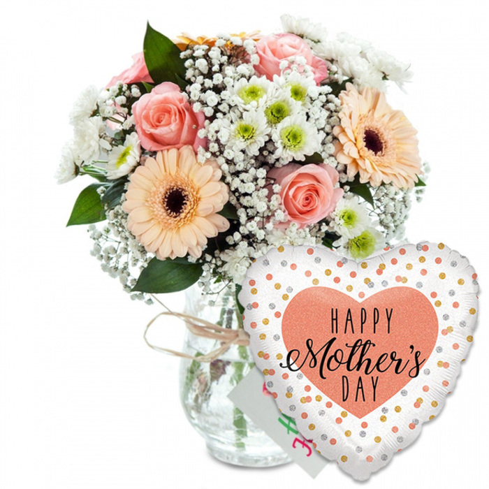 A thoughtfully styled Mother's Day gift ensemble is photographed against a clean white background, making every colour, texture, and reflection stand out with clarity. On the left, a full, dome-shaped bouquet overflows from a clear glass jar-style vase, the stems visible through the water like green brushstrokes. The neck of the vase is encircled by a light brown twine bow, giving the whole piece a subtle rustic charm that feels at home among Clerkenwell's independent studios and cafes. The flowers themselves are a harmonious mix of soft pastels and fresh greens: several peach and blush-pink roses open gracefully, each petal curling outward in a classic spiral, while two or more peach-orange gerbera daisies with deep brown centres provide a jubilant focal point. Around these main blooms, smaller white daisy-type chrysanthemums with pale green centres add both brightness and structure, echoing the fresh, simple beauty of flowers seen in local churchyard gardens. Fine sprays of white gypsophila thread between the larger blooms, giving the arrangement a light, airy finish, as if a soft spring breeze had passed through. Rich green foliage supports and frames the flowers, creating depth and a clear outline against the white backdrop. To the lower right of the bouquet, partly covering the bottom of the vase, a glossy heart-shaped foil balloon leans into the composition. Its white surface is sprinkled with sparkling gold and silver polka dots, catching hints of reflected light. At the centre sits a solid peach heart with the phrase HAPPY Mother's DAY printed inside in contrasting black fonts-bold, simple lettering for HAPPY, and an elegant, flowing script for Mother's DAY. Just behind the balloon's edge, the corner of a small white greeting card is visible, reinforcing the idea of a complete Mother's Day gift ready to be delivered across Clerkenwell, from St John Street to the quiet residential lanes nearby.