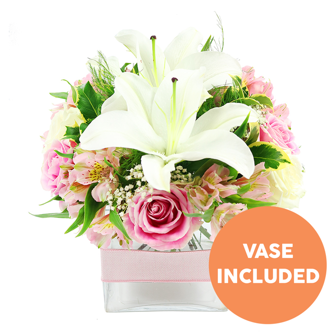 Set against a simple white background, this expertly arranged bouquet looks like it has been styled on a workbench in a Clerkenwell design loft, with every flower placed to create balance and calm. Two large white Oriental lilies rise from the centre, their broad, slightly ruffled petals opening generously like elegant trumpets, each bloom showcasing a cluster of dark reddish-brown stamens and a pale central pistil that draw the eye inward. Nestled closely around these lilies are numerous soft pink roses, ranging from tight, conical buds just beginning to unfurl to fully opened blossoms with layered, velvety petals that create a classic, romantic swirl. Sprays of alstroemeria weave through the roses and lilies, their smaller trumpet-shaped flowers in shades of pink and white, some petals gently brushed with yellow and dotted with fine speckles, introducing a subtle pattern and texture that feels almost hand-illustrated. Light, cloud-like tufts of white baby's breath fill the gaps between the larger blooms, giving the composition an airy, floating quality. Surrounding the entire arrangement is an assortment of fresh greenery - broad, glossy leaves with a rich, deep green tone, slimmer arching blades, and delicate, fern-like fronds - which provide contrast and frame the softer colours. The bouquet sits in a clear square glass vase, its clean geometric lines visible beneath the mass of blooms, stems neatly aligned inside. Around the centre of the vase, a pale pink satin ribbon is wrapped smoothly, adding a gentle decorative band rather than a bow, as if prepared for discreet yet elegant gifting. In the bottom right corner of the image, a bold circular orange badge with white capital letters states VASE INCLUDED, clearly indicating that the container is part of the item. The overall mood is serene and refined, a perfect floral gift for birthdays, anniversaries, or quiet congratulations sent to homes and studios across Clerkenwell and nearby Farringdon.