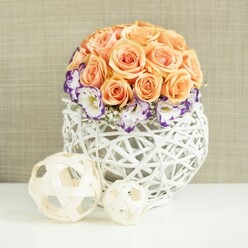 On a clean, white tabletop that could just as easily be a polished counter in a Clerkenwell townhouse, a sophisticated floral centerpiece draws the eye with its calm harmony and sculptural shape. The main focus is a lush, domed cluster of soft peach and apricot roses, densely packed to form a near-perfect sphere of petals. Each rose is fully open, with plush, layered whorls that give a sense of both fullness and tenderness, as though created for a quiet celebration near St John Street. Cradling the base of the rose globe is a graceful ring of lisianthus, their white petals traced with deep violet and purple edges. The colour on the frilled rims adds a gentle drama, reminiscent of hand-painted porcelain, while tiny, cloud-like tufts of white baby's breath are scattered between the blooms to lend lightness and space. The arrangement rests inside a white, bulbous container woven from interlaced wicker strips. This open, basket-like lattice allows light to slip through, casting subtle shadows and underlining the artisan craftsmanship typical of local studios off Clerkenwell Road. In front of the main arrangement, two decorative woven spheres in natural beige tones, one smaller and one slightly larger, sit on the same surface like minimalist sculptures, echoing the texture and shape of the vase. The neutral, softly textured backdrop wraps the scene in warmth, creating a mood of considered elegance and serenity. It feels ideal for a wedding table at a Clerkenwell venue, a refined dinner party, or a styled photoshoot capturing the area's blend of history, design, and understated romance.