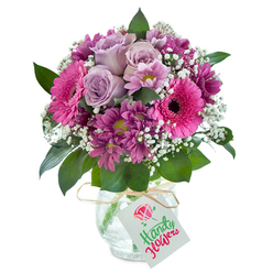 With a distinctive Clerkenwell flair, this lush hand-tied bouquet captures the vibrant energy of the area while retaining a sense of gentle intimacy. Presented in a uniquely textured clear glass vase set against an immaculate white background, the bouquet is elegantly domed, each bloom selected for both visual impact and subtlety. Centered are velvety pale lilac roses, their unfurling petals providing a soft, romantic core redolent of early spring air. These roses are surrounded by ample, wide-faced Gerbera daisies in luminous fuchsia, their striking dark eyes drawing in the gaze much like the inviting storefronts of St John Street. Nestled around the larger flowers, clusters of plum and magenta chrysanthemums offer striking contrast and tactile richness, their daisy-like shapes reminiscent of the creative mosaic found in local design studios. Wisps of delicate white baby's breath flit across the bouquet, softening the overall look and imbuing it with an airy, celebratory feel. The lush, polished green foliage at the base imparts a hint of Clerkenwell Green's leafy charm. The vase's crackled surface catches the light like stained glass, and a tightly knotted natural raffia ribbon, accented with a cheerful birthday tag decorated in red and green, completes the ensemble. The result is a visually stunning, emotionally evocative creation perfect for marking milestones, birthdays, or simply bringing a little urban joy to someone special across Clerkenwell.
