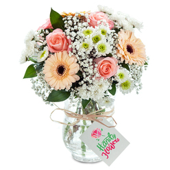Imagine stepping into a Clerkenwell morning, where sunlight streams through loft windows and the day begins with a gentle sense of promise. This captivating bouquet captures that very mood, intertwining soft peach gerberas and blush-pink roses that unfurl in delicate, embracing layers. Airy sprigs of baby's breath float between the main blooms-innocent and light, much like the clouds drifting above St James's Churchyard nearby. White chrysanthemums, fresh and rounded, nestle throughout, each flirting with a hint of chartreuse at their center, reminiscent of new beginnings and local gardens after rain. The tactile richness is striking; fingers would glide over silky, cooling petals and brush against feathery gypsophila, all anchored by a vase of clear, subtly textured glass. Gleaming stems are visible through the water, exuding an unmistakable just-picked vibe. A neat bow of rustic raffia sits at the neck of the vase, evoking the relaxed yet cultured charm found in Clerkenwell's coffee shops along Clerkenwell Green. Suspended from the ribbon is an unmarked rectangular white tag, ready to bear a heartfelt message. This bouquet feels like a quiet celebration-for birthdays, anniversaries, or sending comfort-a gentle floral embrace grounded in Clerkenwell's stylish heritage, artfully arranged for delivery across the neighbourhood.