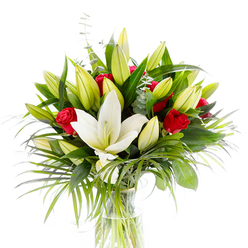 A luminous arrangement whose first note is the sculptural white oriental lily set forward and open, its creamy petals unfurling with serene confidence, revealing delicate pale-yellow stamens and a slender green pistil. Encircling that focal bloom are numerous lily buds in fresh lime and soft yellow tones - a chorus of futures waiting to bloom - while vivid red rosebuds peek through, their velvety petals still curled in on themselves like whispered promises. The greenery composes the scene: dense dark green lily leaves give weight and form, long feathered palm fronds in an emerald hue lend graceful motion, and small stems of silvered eucalyptus add a cool, dusty contrast and delicate scent line. A simple clear glass vase holds the ensemble, the submerged stems visible in clean water and grounding the arrangement in natural simplicity. Against the bright white background the contrasts sing: ivory and ruby, deep green and soft silver, light and shadow creating subtle depth. One can almost sense the fragrance - a soft powderiness from the lilies with the warmer, richer trace of roses - and the image carries an emotional warmth suited to celebratory gestures, a romantic gift, or a refined sympathy piece. For a local touch, this is the kind of bouquet a Clerkenwell resident might choose for a dinner at a townhouse near St. John's Square or to send to friends across Leather Lane; it reads as thoughtful, professional and perfectly composed for urban floral gifting.