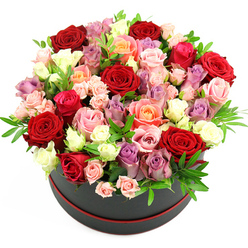 A sumptuous round hatbox arrangement brimming with roses is shown in close detail, as if placed on a spotless studio table somewhere off St John Street in Clerkenwell. The container is a sleek, cylindrical box in deep black with a fine red trim circling its base, a subtle flourish that hints at a premium flower gift prepared with care. Roses cascade over the edges, creating a lush, domed canopy of petals that feels full, generous, and expertly constructed. Deep red roses, velvety and rich like a glass of Merlot sipped in a cosy Clerkenwell bar, sit at the heart of the bouquet and draw the eye with their intensity. Around them, lighter tones unfold: blush pink and fuchsia blooms bring soft romance and playfulness, while peach and apricot roses add warmth, like late-afternoon sun across nearby Clerkenwell Green. Creamy white and ivory roses introduce a sense of purity and calm, their smooth petals catching the light with a gentle sheen. Sprinkled among them are elegant lavender and muted purple roses, giving the composition a refined, contemporary twist that suits the artistic studios of the area. The roses vary in size and stage of bloom: open rosettes with layered, ruffled petals nestle alongside smaller spray roses and tight buds, adding fine detail and a sense of natural progression. Slender pieces of fresh green foliage, perhaps eucalyptus or ruscus, thread through the arrangement, breaking up the colour with cool, refreshing greens that suggest morning market freshness. Set against a clean white backdrop, this hatbox bouquet exudes romance and celebration, ideal for an anniversary, a special birthday, or a heartfelt thank-you delivered across Clerkenwell and the surrounding central London neighbourhoods.